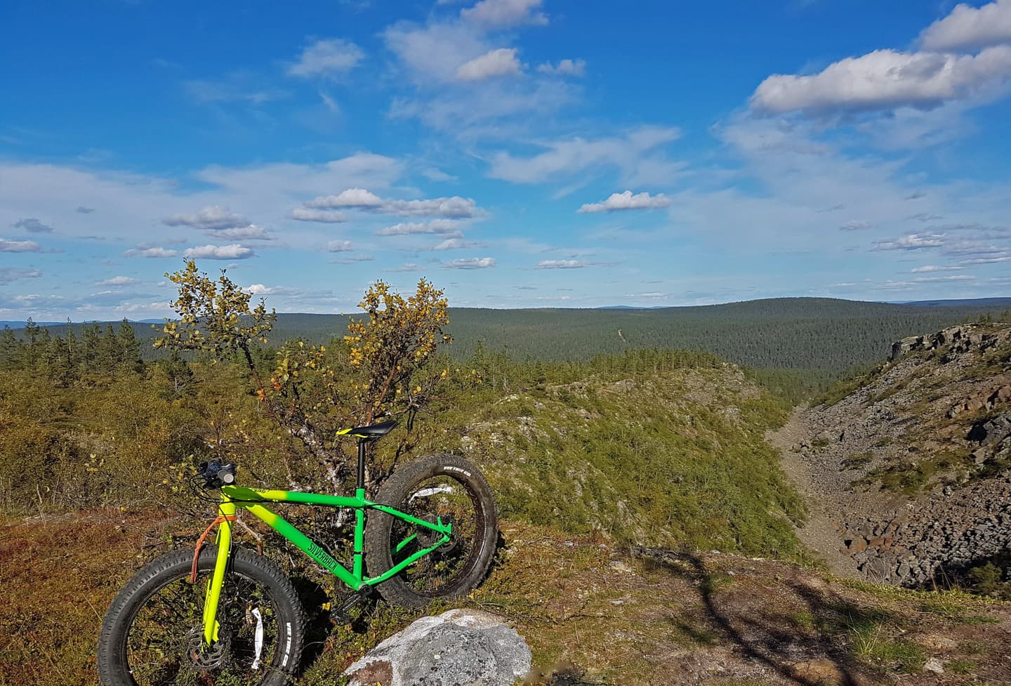 Bild: Juho Niemelä Det finns en mountainbike vid kanten av en brant ravin. Vid ravinens kant finns några fjällbjörkar. På himlen finns det vackert väder-moln och solen skiner.