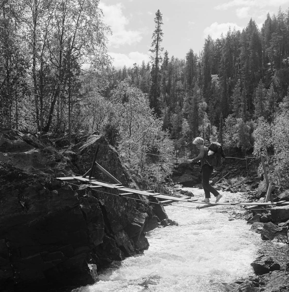 Kuva: Matti Poutvaara Vanha mustavalkoinen valokuva. Retkeilijä ylittämässä koskea riippusillalla.