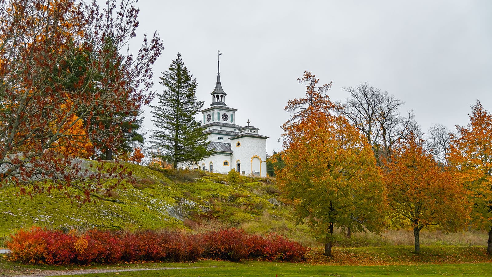 Bild: Jasmin Yuchun En kyrka omgiven av höstträd. Träden är fulla av färgglada löv i nyanser av gult, orange och rött. Den molniga himlen i bakgrunden förstärker höstatmosfären.