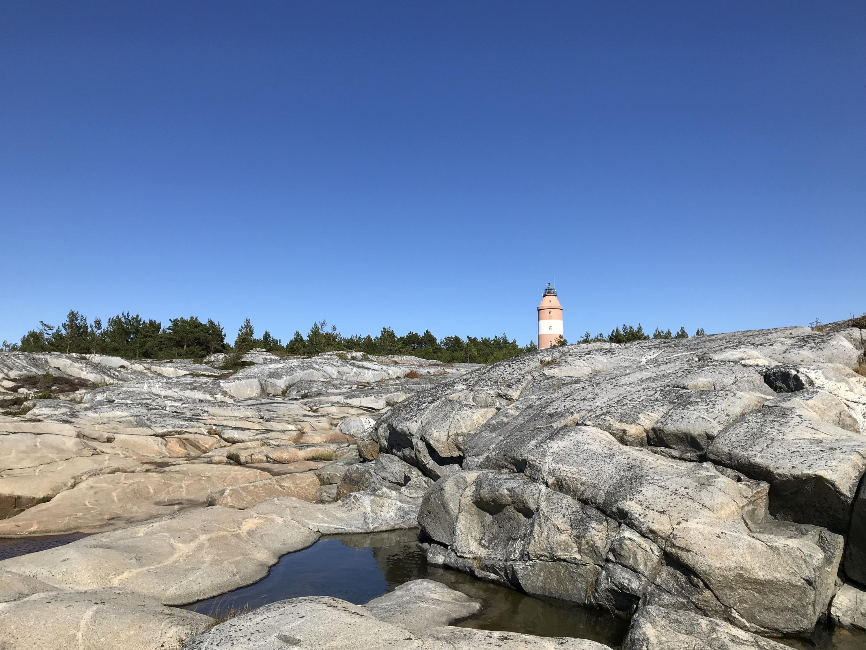 Image: Elina Kuusniemi A rocky island. In the distance, a small red and white striped lighthouse is visible.