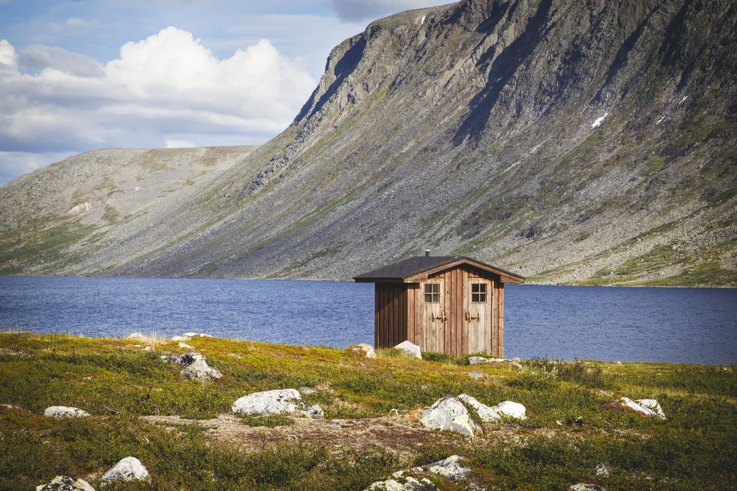 Bild: Veli-Pekka Heikkilä / Vastavalo En liten byggnad med två dörrar vid stranden på sommaren. På andra sidan sjön reser sig ett fjäll.