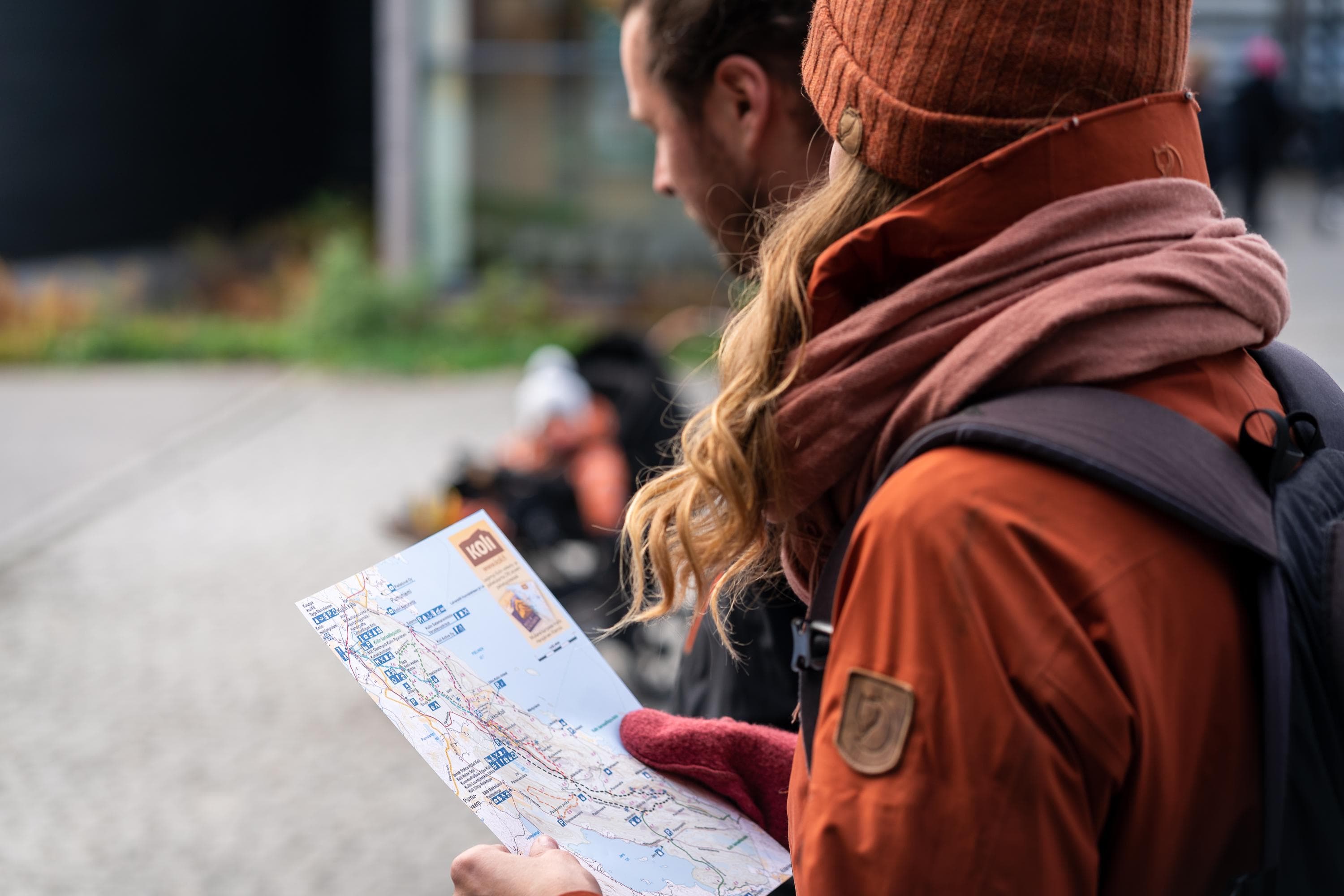 Kuva: Mitja Piipponen Edustalla nainen punaisessa pipossa ja takissa reppu selässä lukee karttaa. Vieressä on mies ja taustalla näkyy lapsi rattaissa. Taaimmaisena näkyy rakennus.