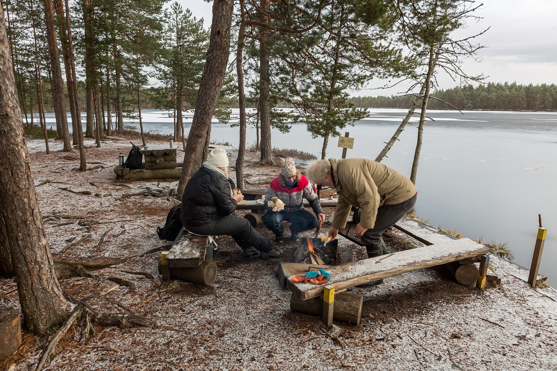 Image: Jari Kostet Kolme retkeilijää rannan tulentekopaikalla. Yksi lisää polttopuuta ja kaksi pitelee eväitä käsissään. Järvi on osin jäässä ja maassa ja penkeillä on hieman lunta.
