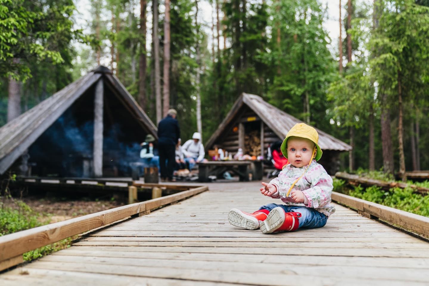 Bild: Jaakko Posti Ett litet barn sitter på den tillgängliga spångade leden. I bakgrunden finns vuxna vid eldstaden.