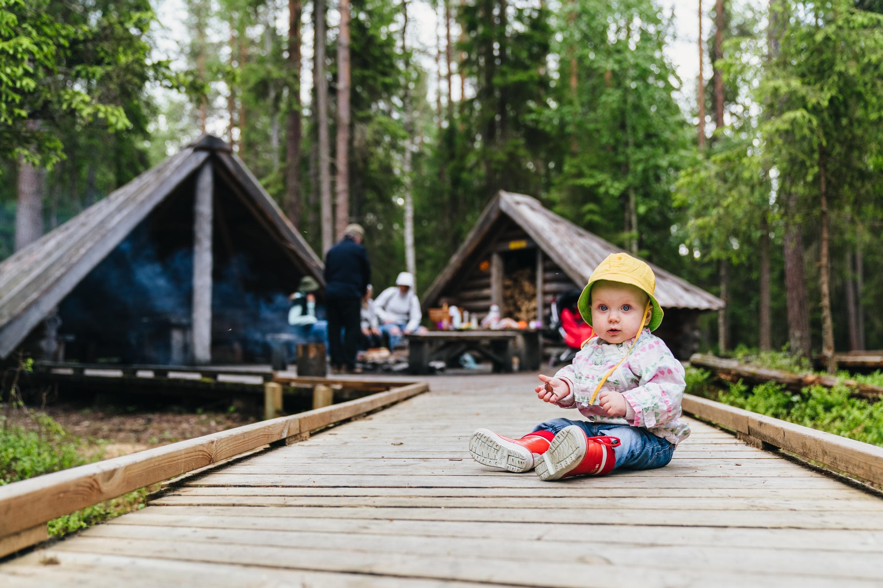 Kuva: Jaakko Posti Pieni lapsi istuu esteettömällä lankkupolulla. Taustalla tulentekopaikalla aikuisia.