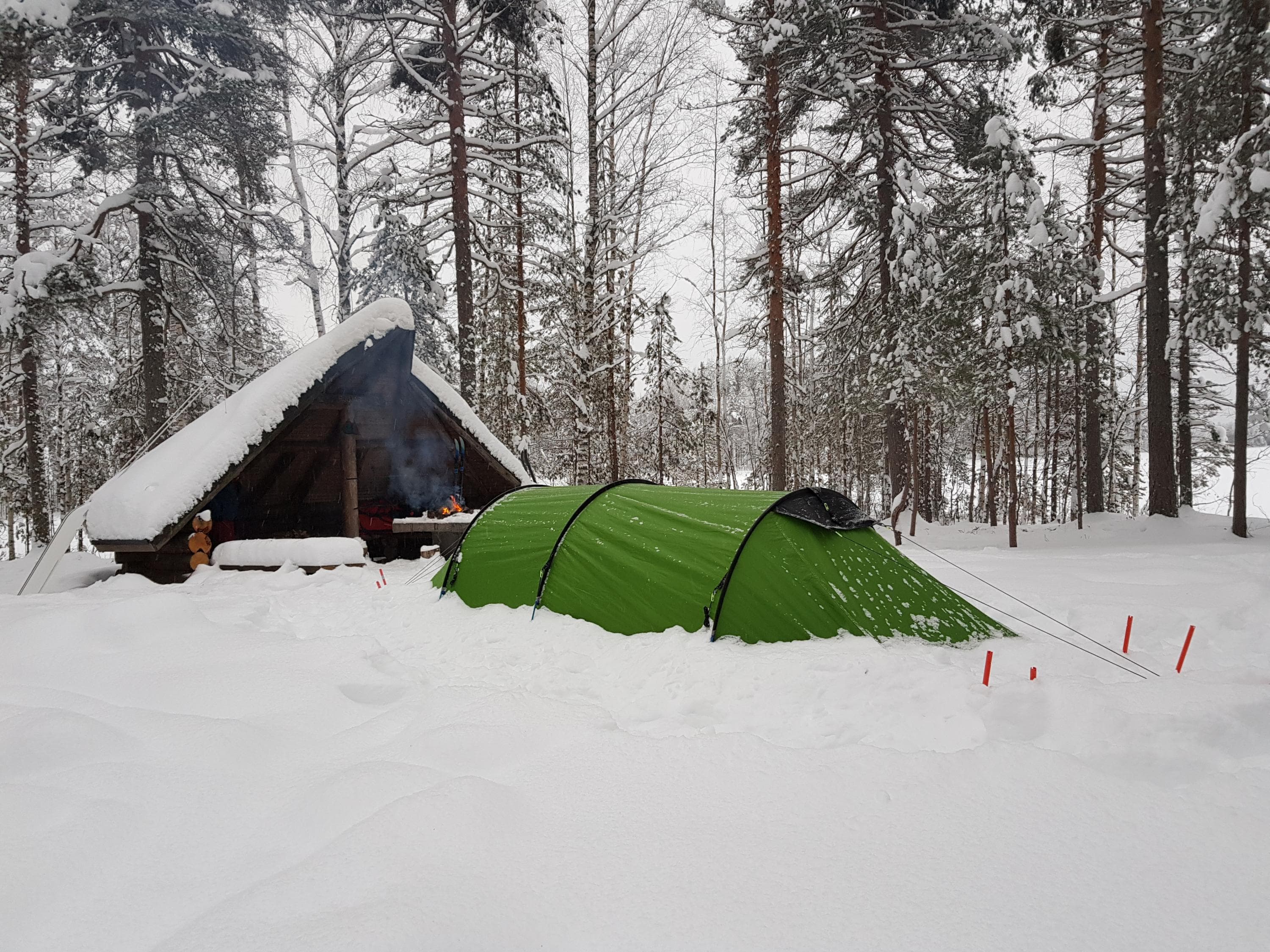 Bild: Sanna Eskelinen / Metsähallitus Kuva lumisesta Harjujärven puolikodasta, jonka viereen on pystytetty vihreä tunneliteltta syvään lumeen. Nuotiossa on tuli.