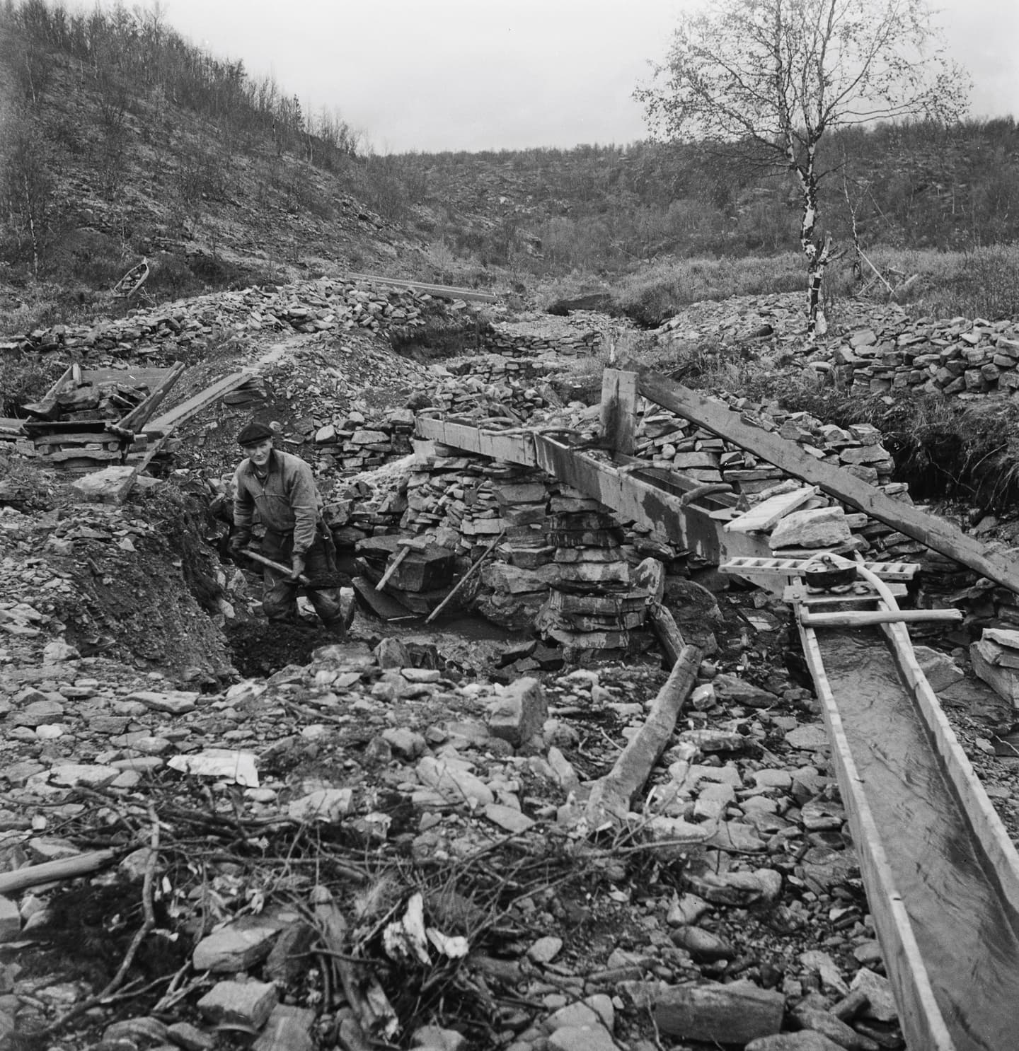 Kuva: Vesa Mäkinen / Museovirasto, Historiallinen kuvakokoelma Mustavalkoisessa kuvassa näkyy kullanhuuhdonta ränni louhikkoisessa maastossa. Kivien keskellä mies lapioi soraa ränniin. Taustalla näkyy lehdettömiä koivuja ja tuntureita.