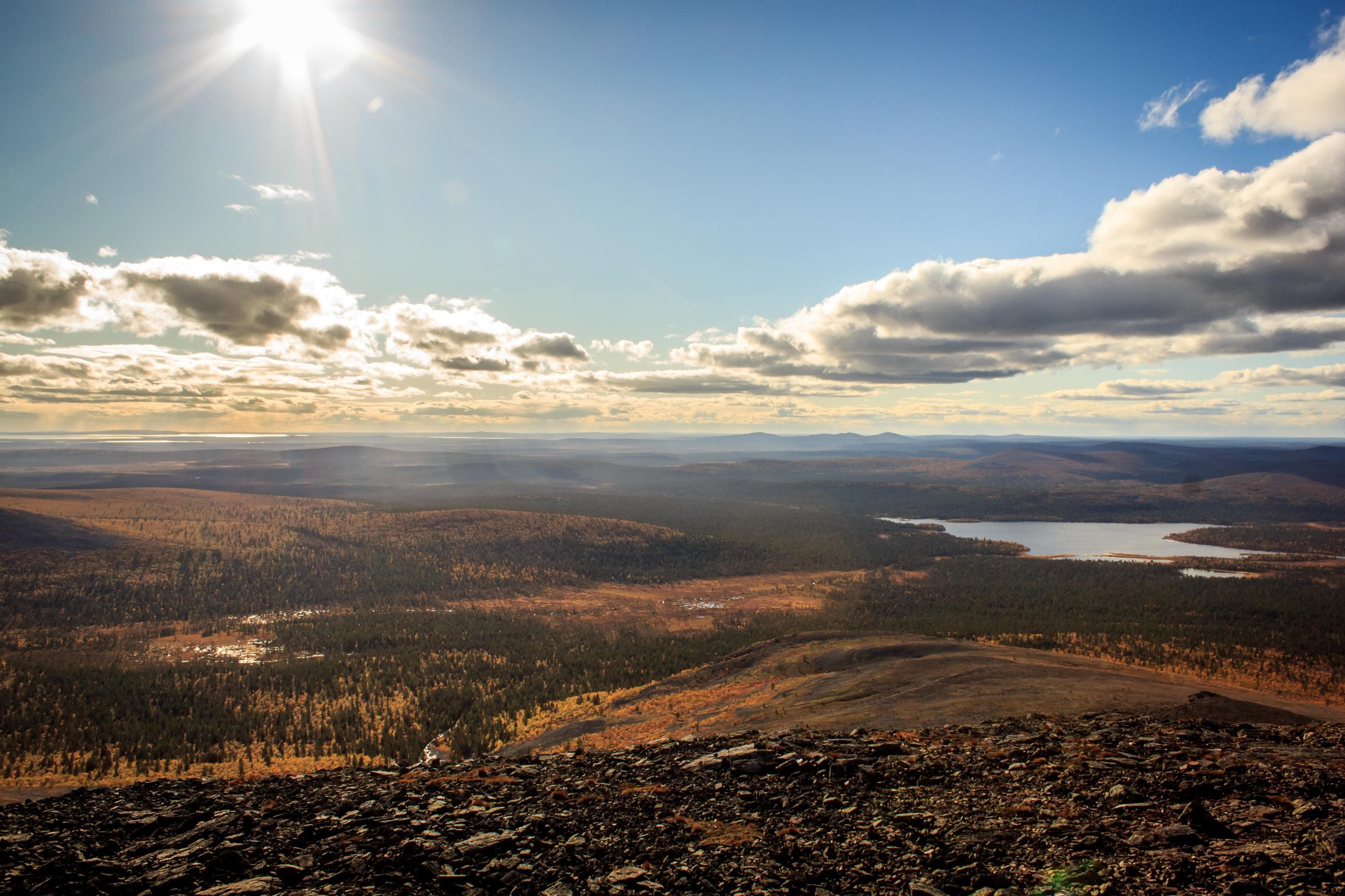 Bild: Juha Kaipainen Bilden togs en solig dag från toppen av ett högt fjäll. Fjällområdet är vidsträckt, och i fjärran kan man se en sjö.