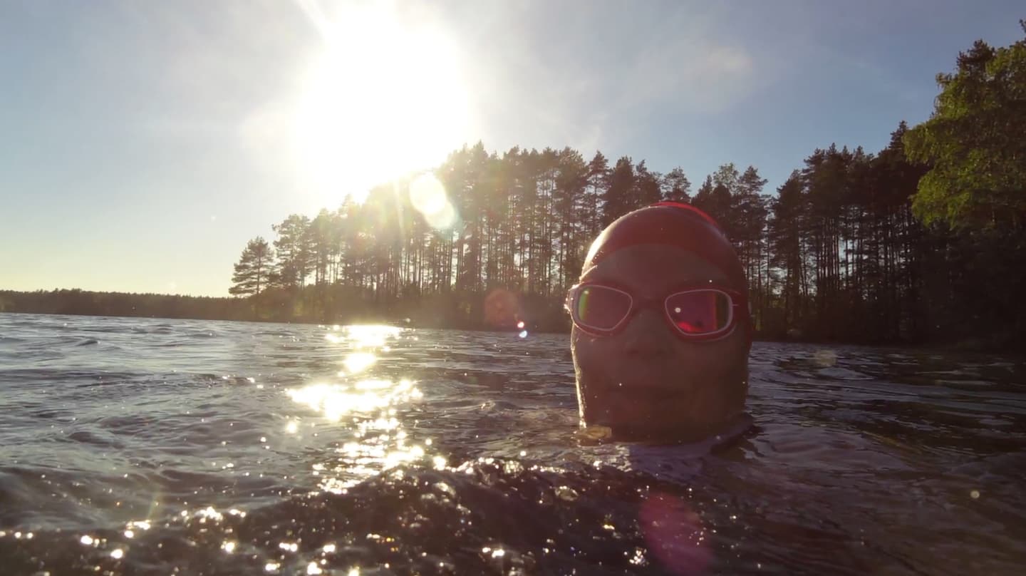 Kove: Päivi Pälvimäki A swimmer standing in a pond with water to his neck, the swimmer is dressed in swimming goggles and a swim cap. A coniferous forest in the background.