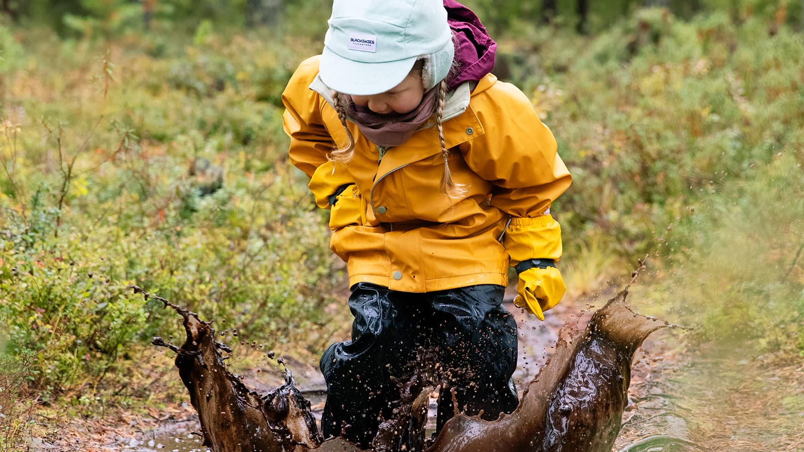 Kuva: Katri Lehtola / Keksi Lapsi hyppii kuralätäkössä sadevarusteissa metsäaukealla.
