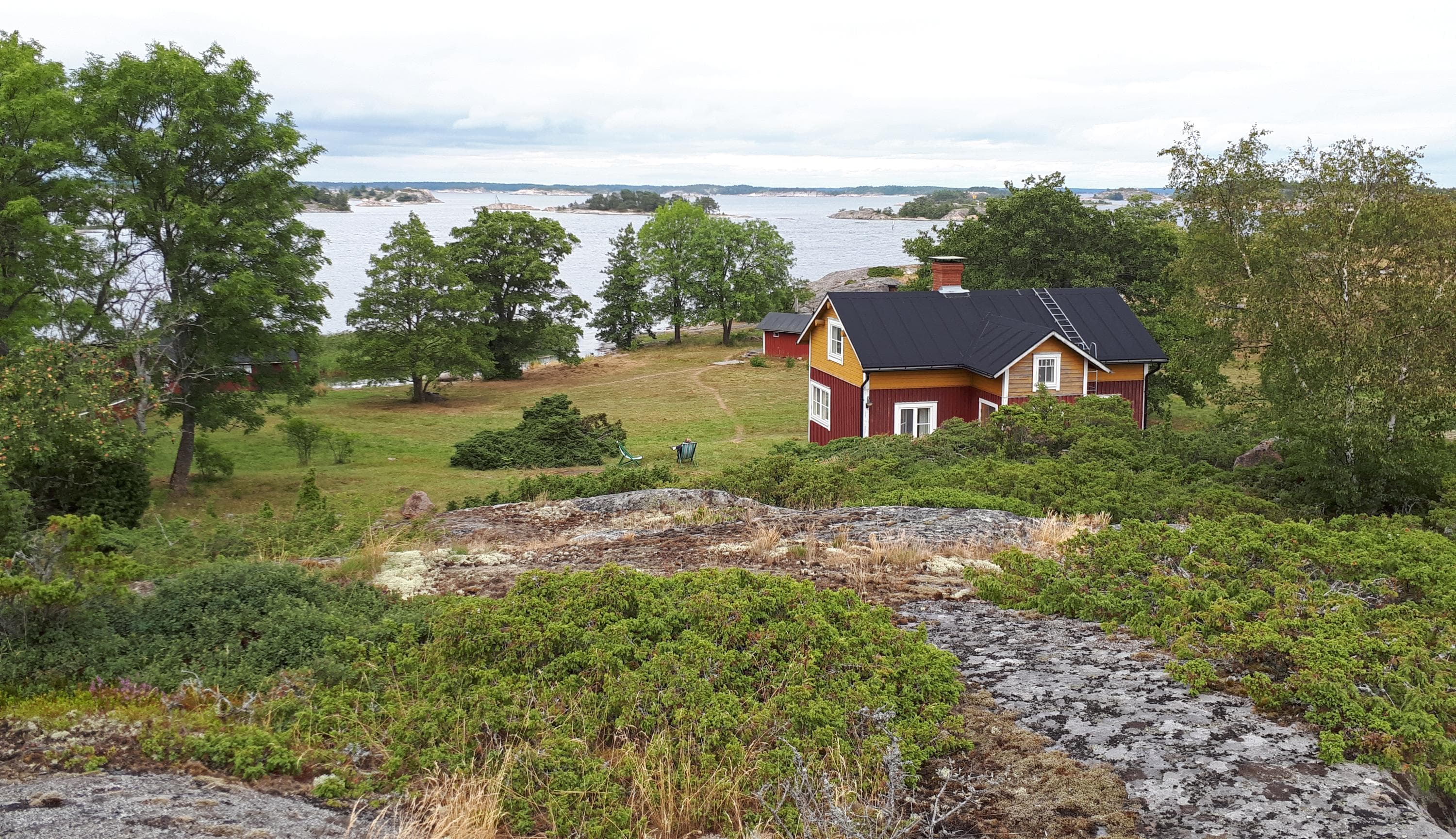Image: Jussi Helimäki A red-yellow wooden house under a rock in the yard surrounded by trees. Sea and islands in the background.