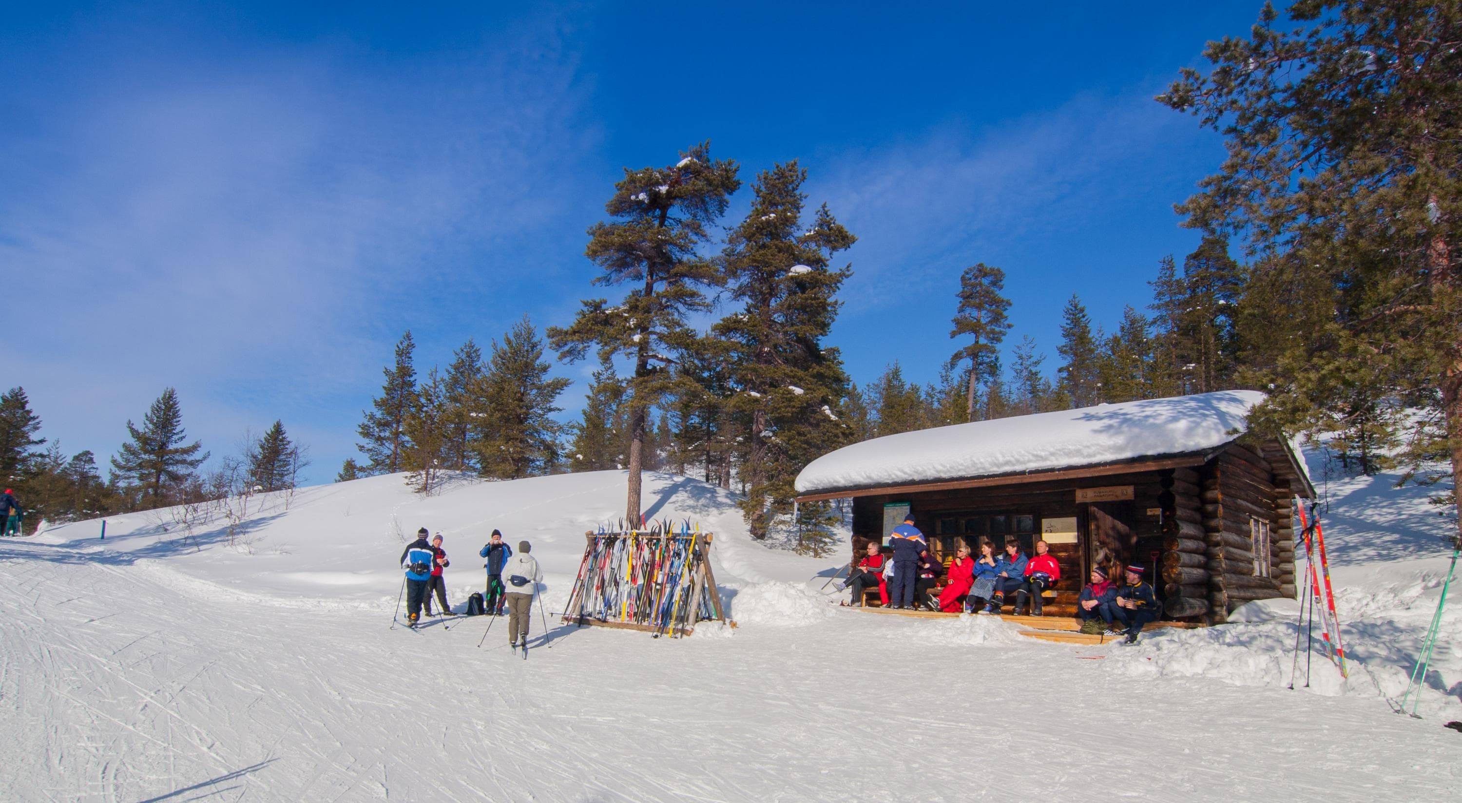 Image: Pertti Turunen / Metsähallitus Hiihtoladun varrella olevan mökin terassilla istuu ihmisiä auringon paisteessa. Mökin edessä hiihtoladulla seisoo muutama hiihtäjä.