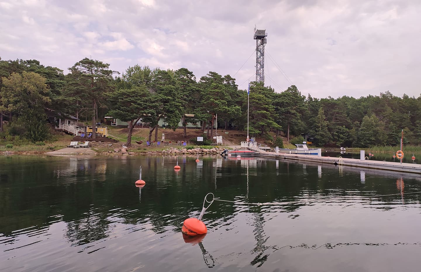 Image: Jussi Helimäki I en lugn havsvik, bojar, en brygga och en strand med byggnader, en skog och ett högt torn i bakgrunden.