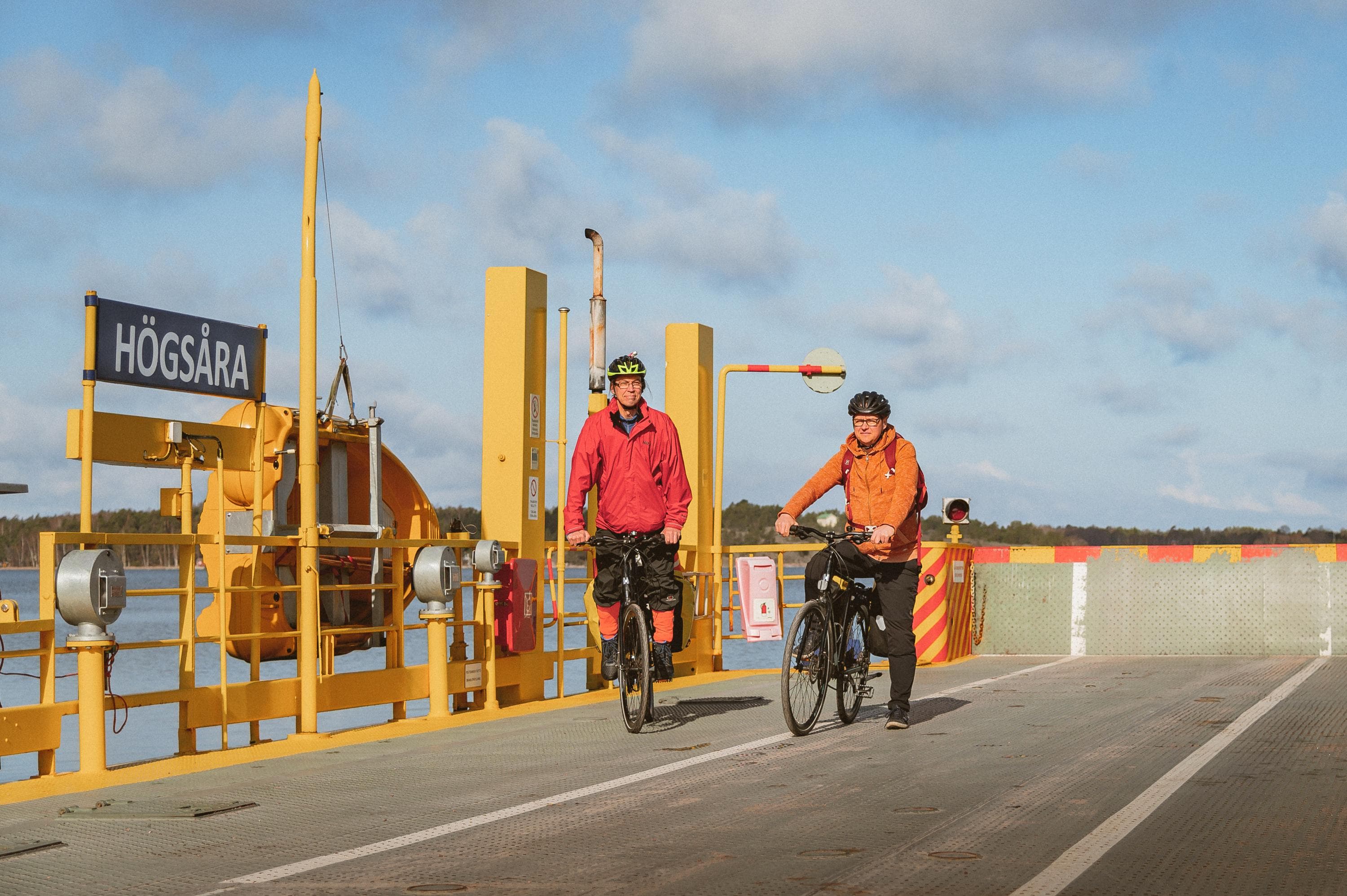Image: Jasmin Yuchun Two cyclists wearing helmets are leaving a ferry, where a sign reads "Högsåra." The ferry is otherwise empty, and the background shows a clear sky with a few scattered clouds.