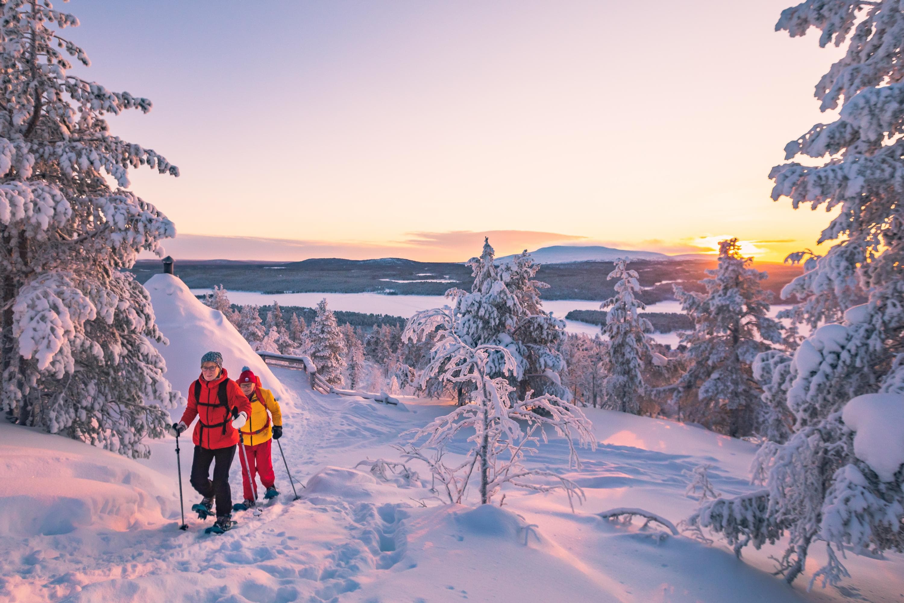 Kuva: Rami Valonen Kaksi retkeilijää nousemassa lumikengillä vaaralle. Ympärillä lumisia mäntyjä ja taustalla horisontissa tuntureita. Aurinko paistaa hyvin matalalta ja se värjää maiseman punertavaksi.