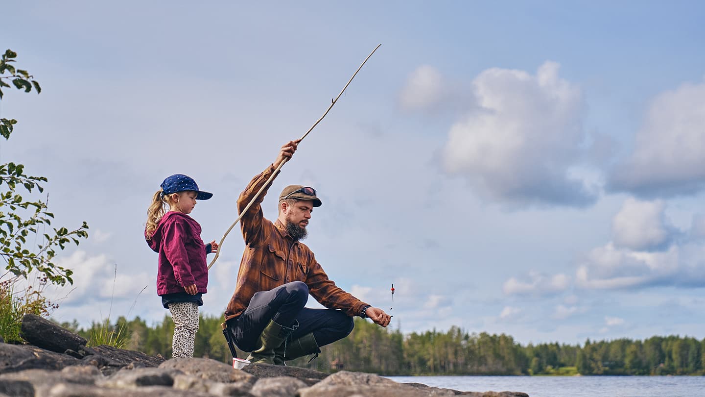 Lapsi seisoo onkivapa kädessä, vanhempi istuu kyykyssä vieressä ja pitelee vapaa ja matoa.