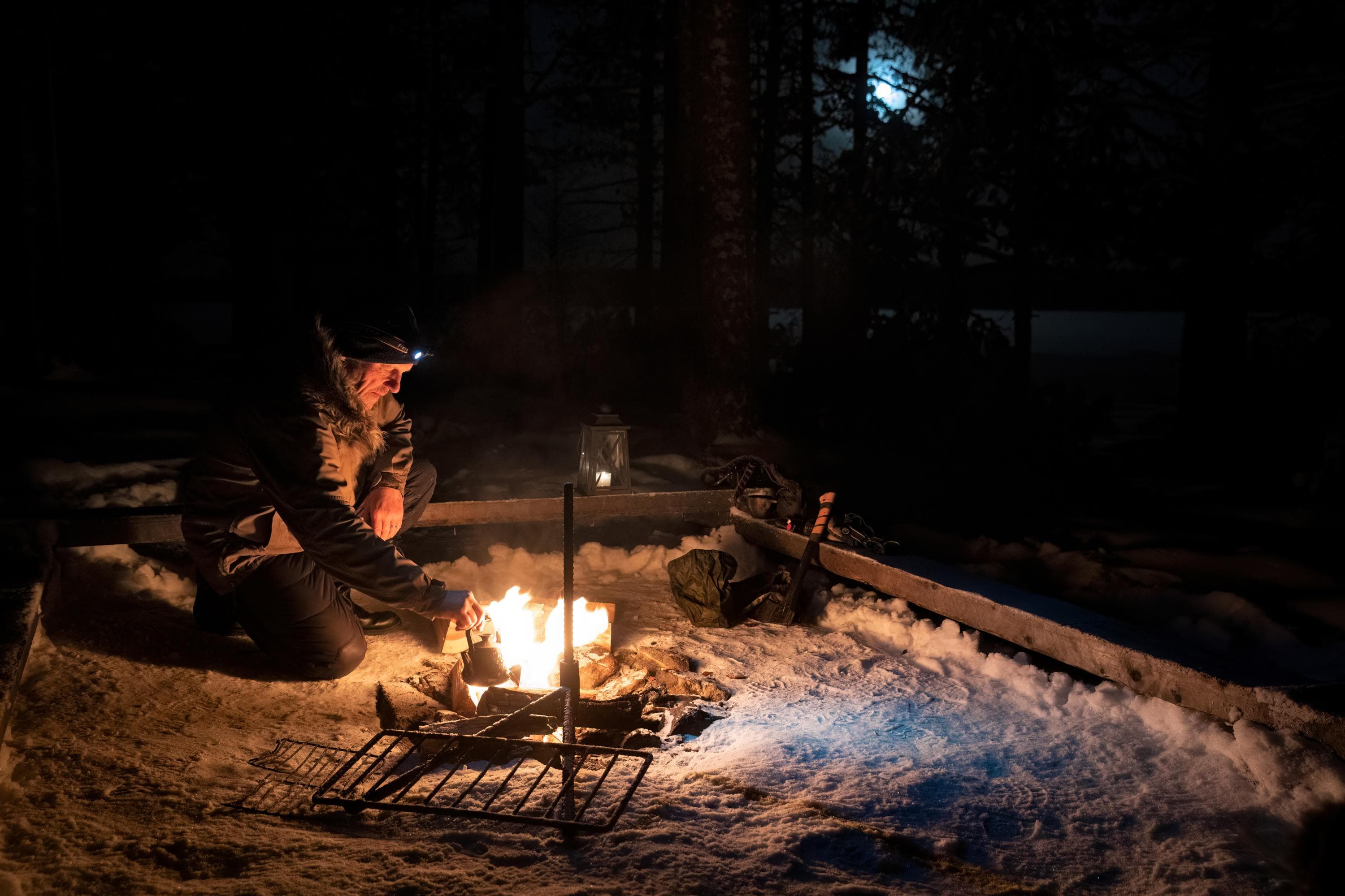 Bild: Katri Lehtola En person sitter vid en snötäckt eldstad i mörkret och lyfter en kaffepanna från elden. Hen har en anorak och en pannlampa. Månen lyser mellan träden i bakgrunden.