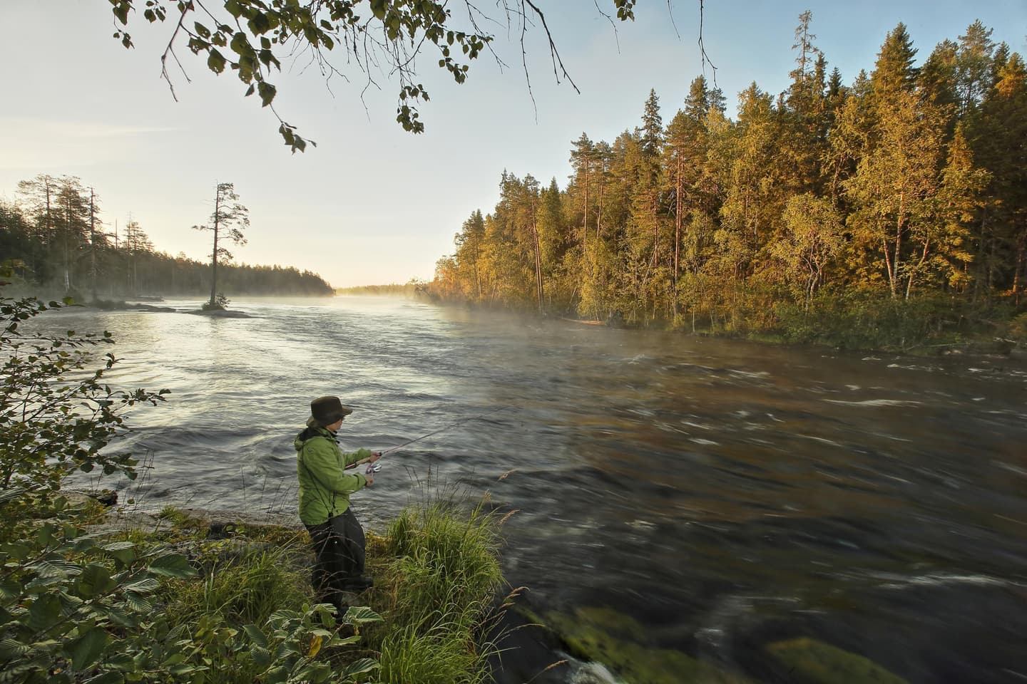 Bild: Ismo Pekkarinen En vandrare fiskar från stranden vid Ruunaa vandringsområde. I bakgrunden höstlig skog.
