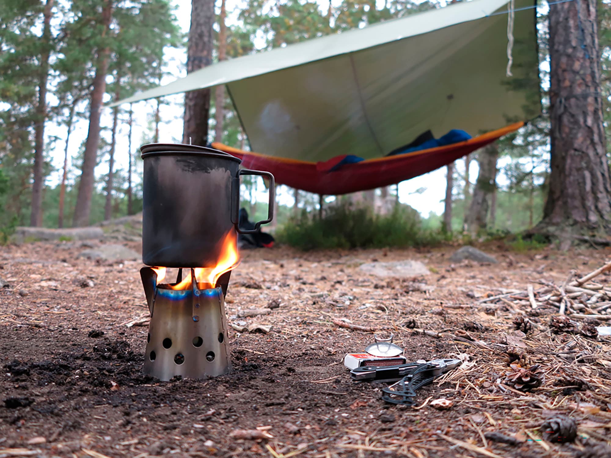 Kuva: Suomen Latu / Juhani Metsäpelto Etualalla risukeitin, jossa ruoka valmistuu. Taustalla metsässä kahden puun väliin on ripustettu riippumatto ja sen yläpuolella on pressukatos.
