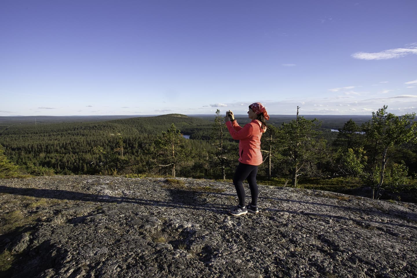 Bild: Minna Koramo En vandrare med en mobiltelefon i handen på toppen av fjället. I bakgrunden en klar himmel, skog, en annan fjälltopp och sjöar.