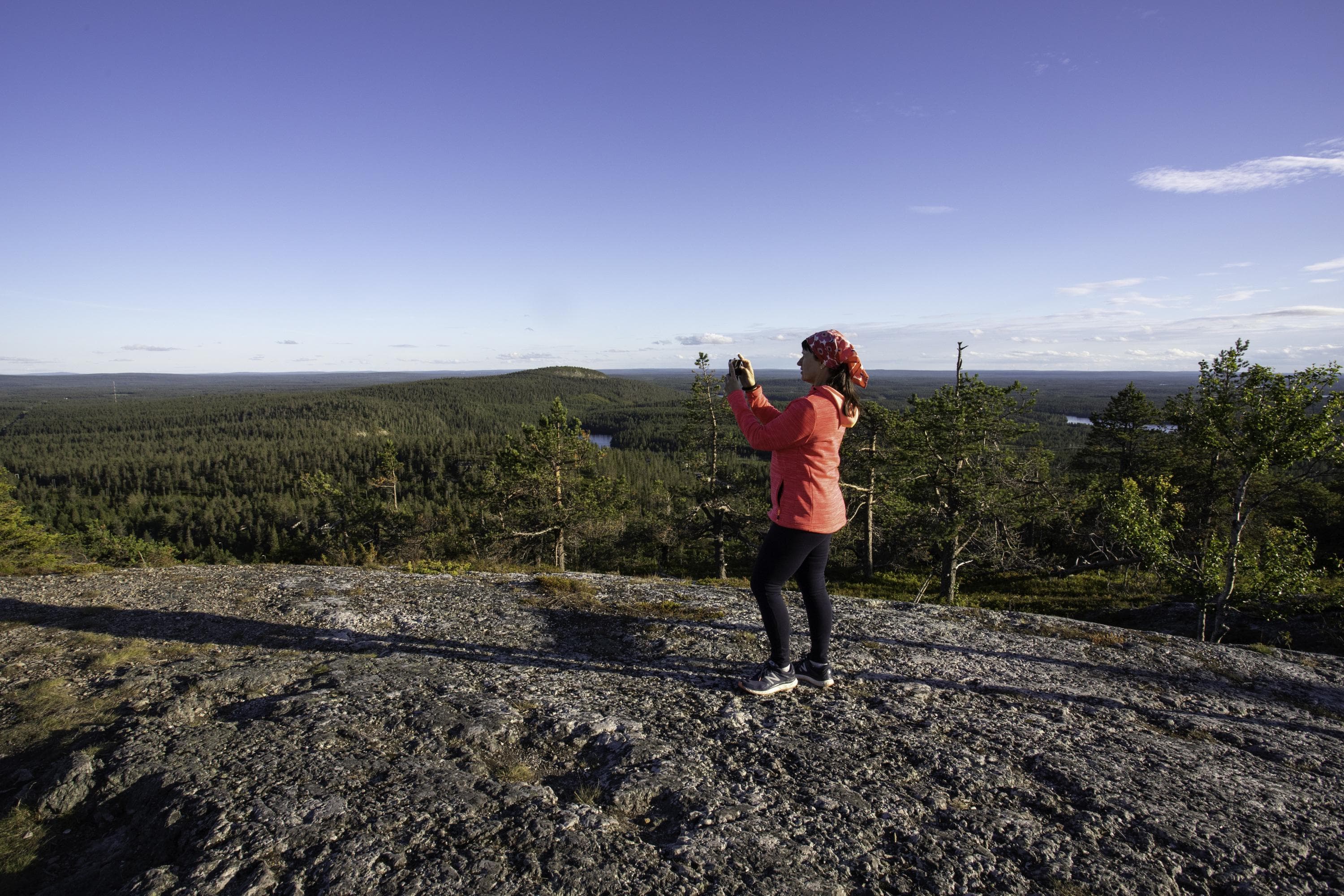 Bild: Minna Koramo Retkeilijä kännykkä kädessä tunturin huipulla. Taustalla poutataivas, metsää, toinen tunturin huippu ja järviä.