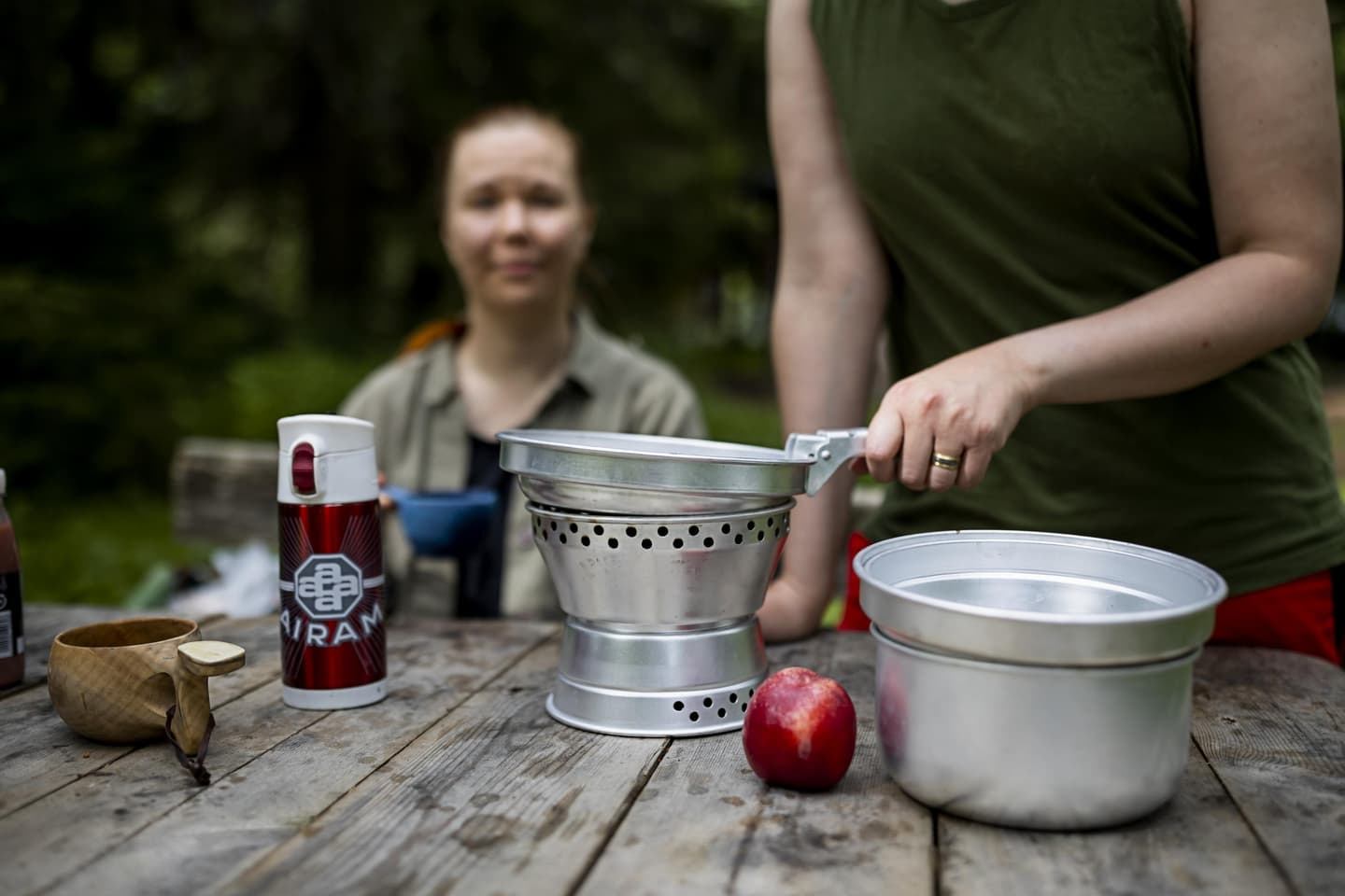 Kuva: Maarit Vaahteranoksa Retkeilijä paistaa retkikeittimen pannulla ruokaa. Pöydällä on kuksa, termospullo ja retkikeittimen kattila. Taustalla toinen retkeilijä seuraa vierestä ruoanlaittoa.