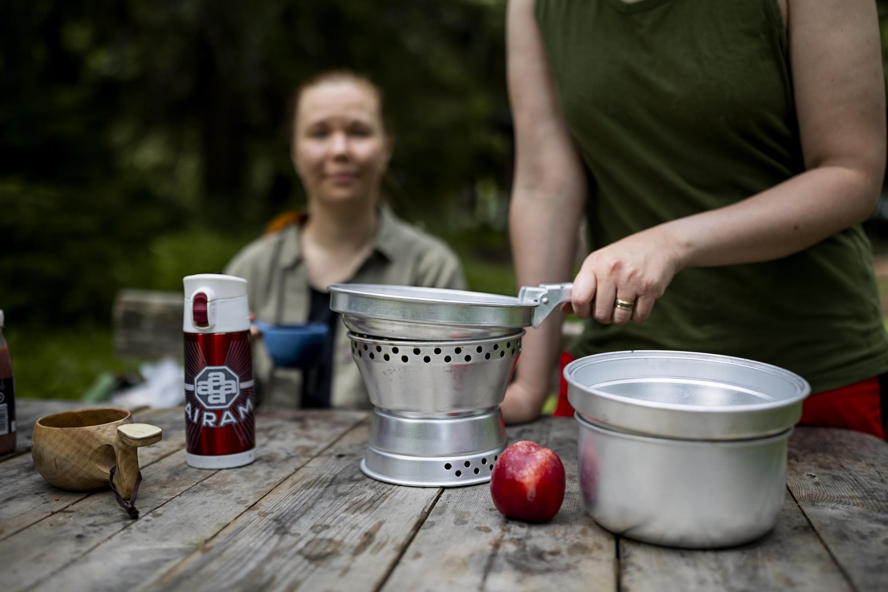 Kuva: Maarit Vaahteranoksa Retkeilijä paistaa retkikeittimen pannulla ruokaa. Pöydällä on kuksa, termospullo ja retkikeittimen kattila. Taustalla toinen retkeilijä seuraa vierestä ruoanlaittoa.