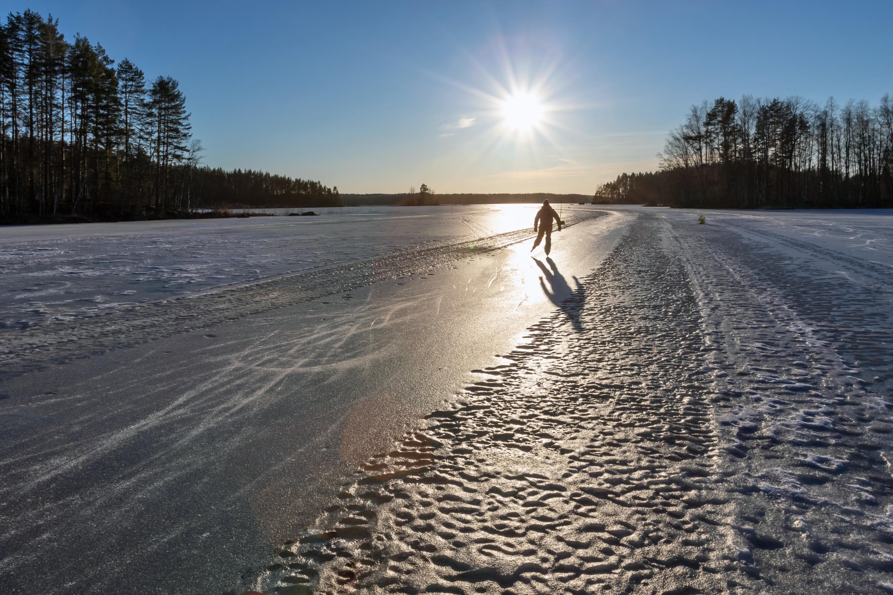 Kuva: Timo Nieminen Retkiluistelija luistelee järven jäälle auratulla luisteluradalla. Aurinko paistaa.