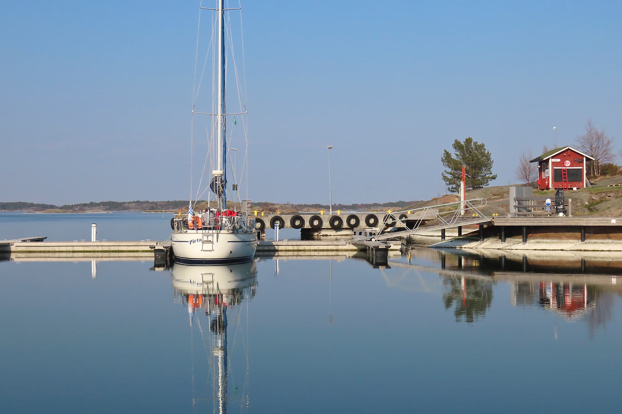 Kuva: Jarmo Vehkakoski Valkea purjevene kiinnitettynä puiseen laituriin. Rannalla kalliota ja punainen rakennus.