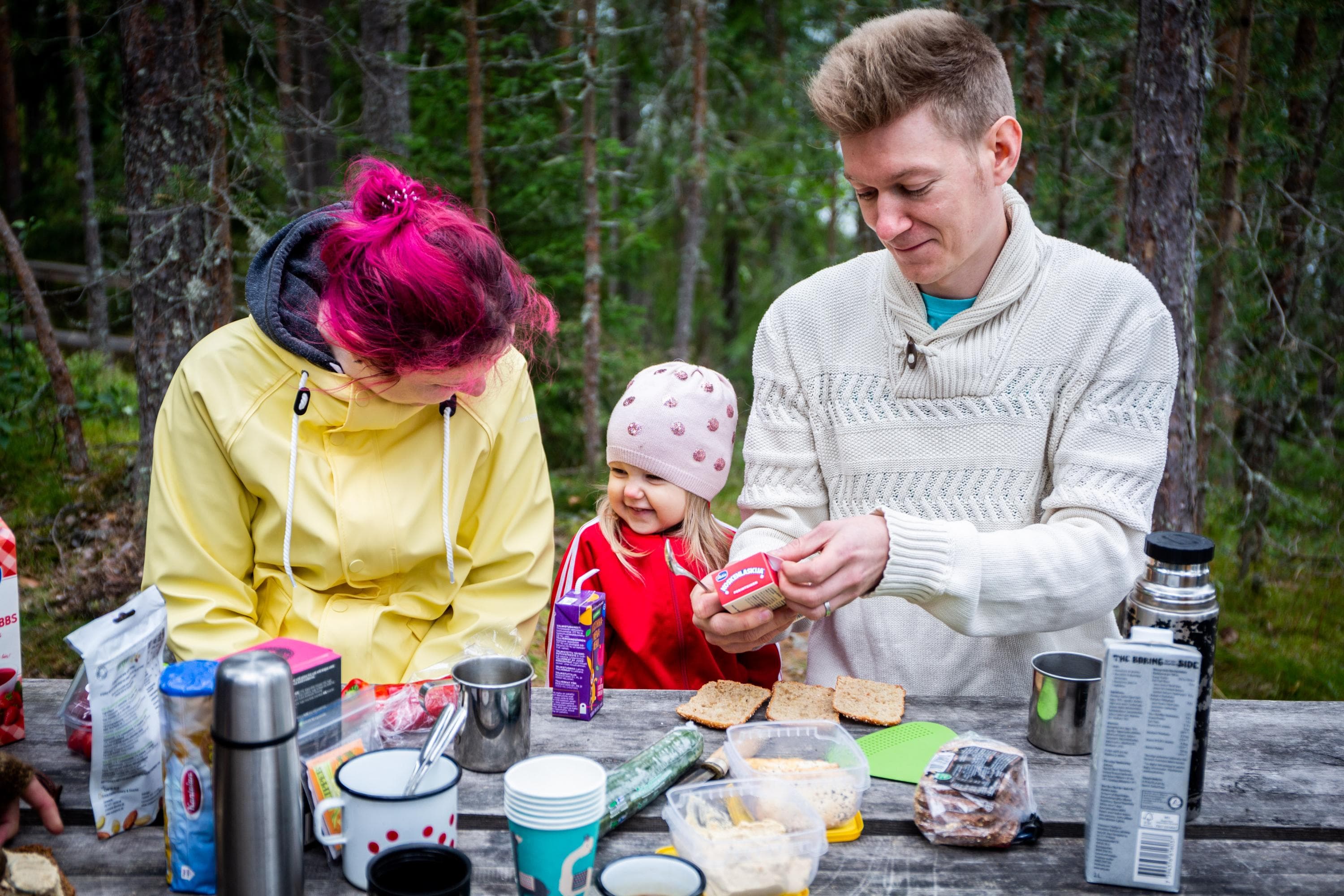 Kuva: Saara Lavi Kaksi retkeilijää ja pieni lapsi syömässä eväitä. Taustalla kesäistä metsää.