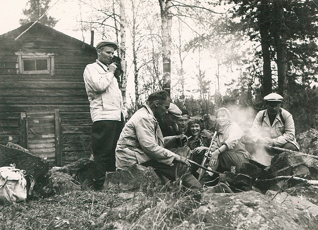 Kuva: Sirkka Saastamoinen Vanha mustavalkovalokuva, jossa ihmiset ovat kokooontuneet nuotion ääreen. Taustalla näkyy hirsirakennus.