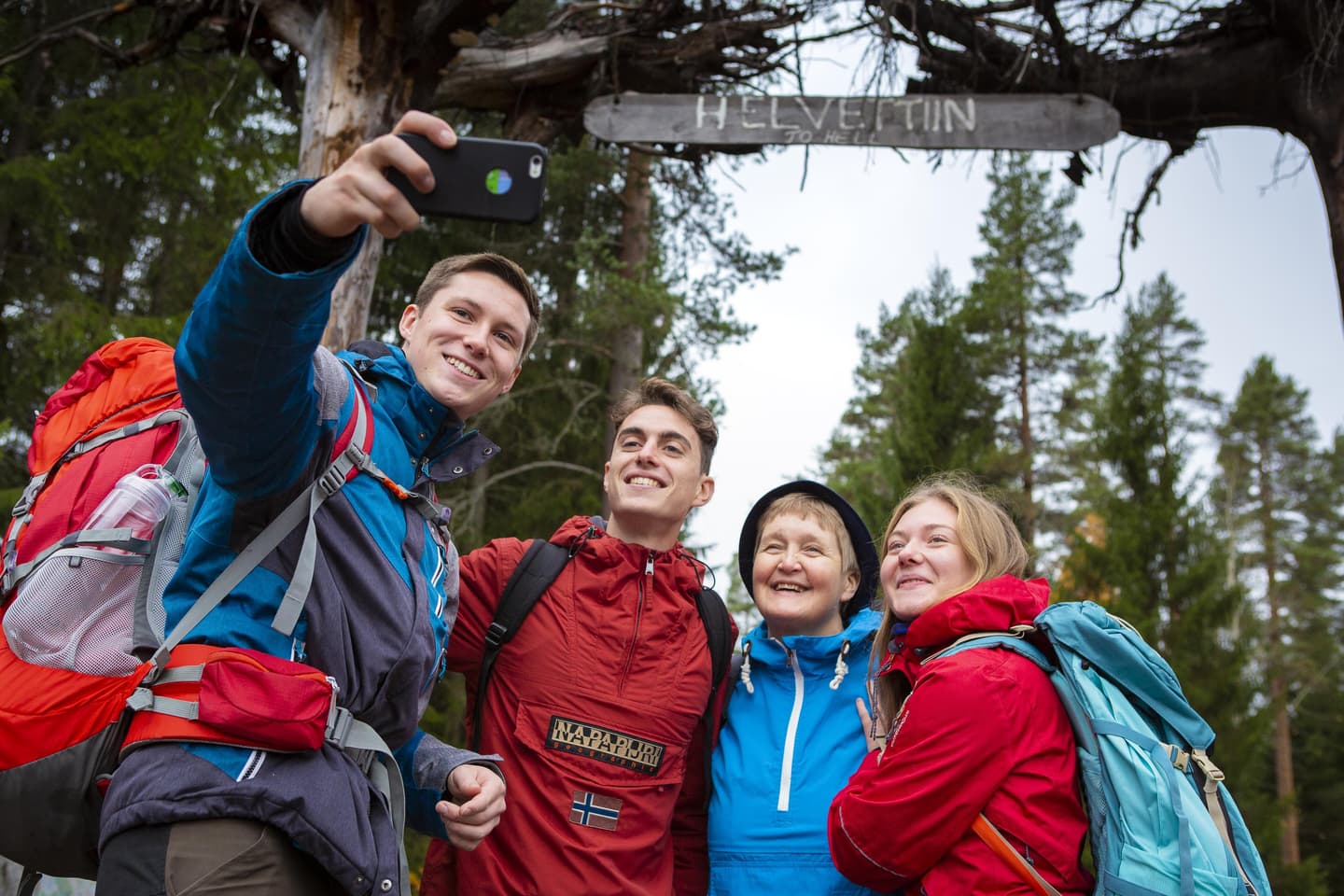Image: Salla Penttilä Four hikers have gathered to take a group photo with a mobile phone. Above them, there is a sign that reads Helvettiin - To hell.