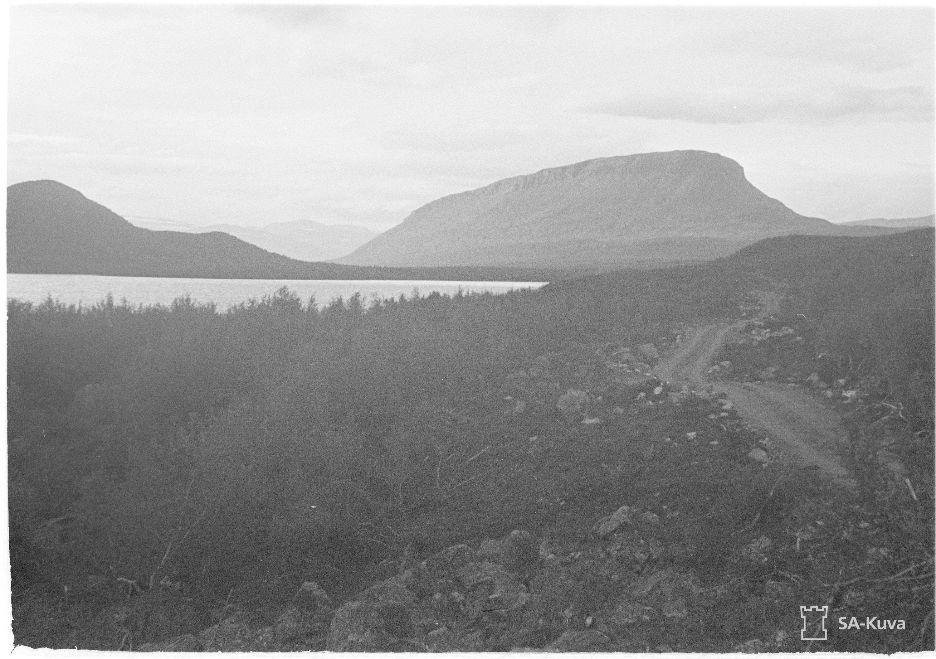 Kuva: SA-kuva-arkisto Mustavalkea SA kuva-arkiston kuva, jossa Saanatunturi ja Pikku-Malla kohoavat Kilpisjärven takana. Etualalla Käsivarrentie ajanmukaisessa hiekkatie muodossa.