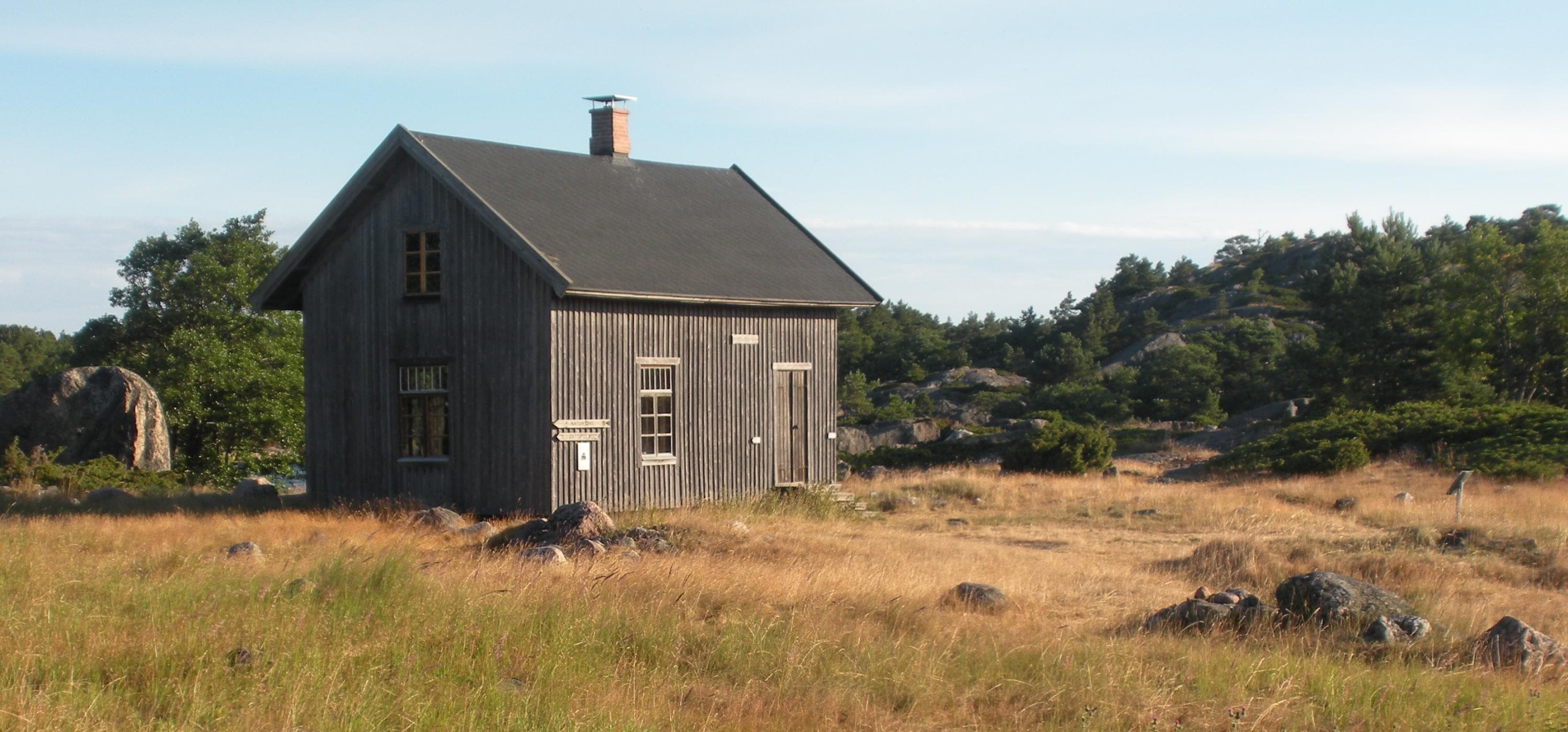 Image: Sanna-Mari Rivasto Grey wooden house in a grassy and stony courtyard, behind the forest.