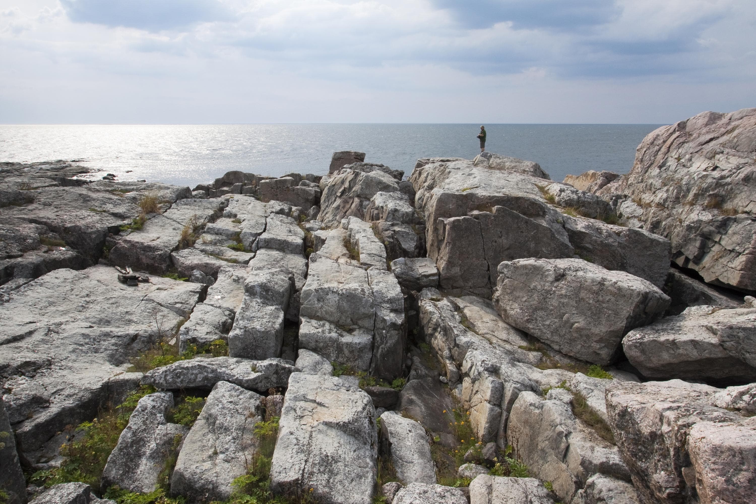 Kuva: Timo Nieminen Kivikkoista merenrantaa. Kaukana kalliolla seisoo ihminen ja katsoo merelle.