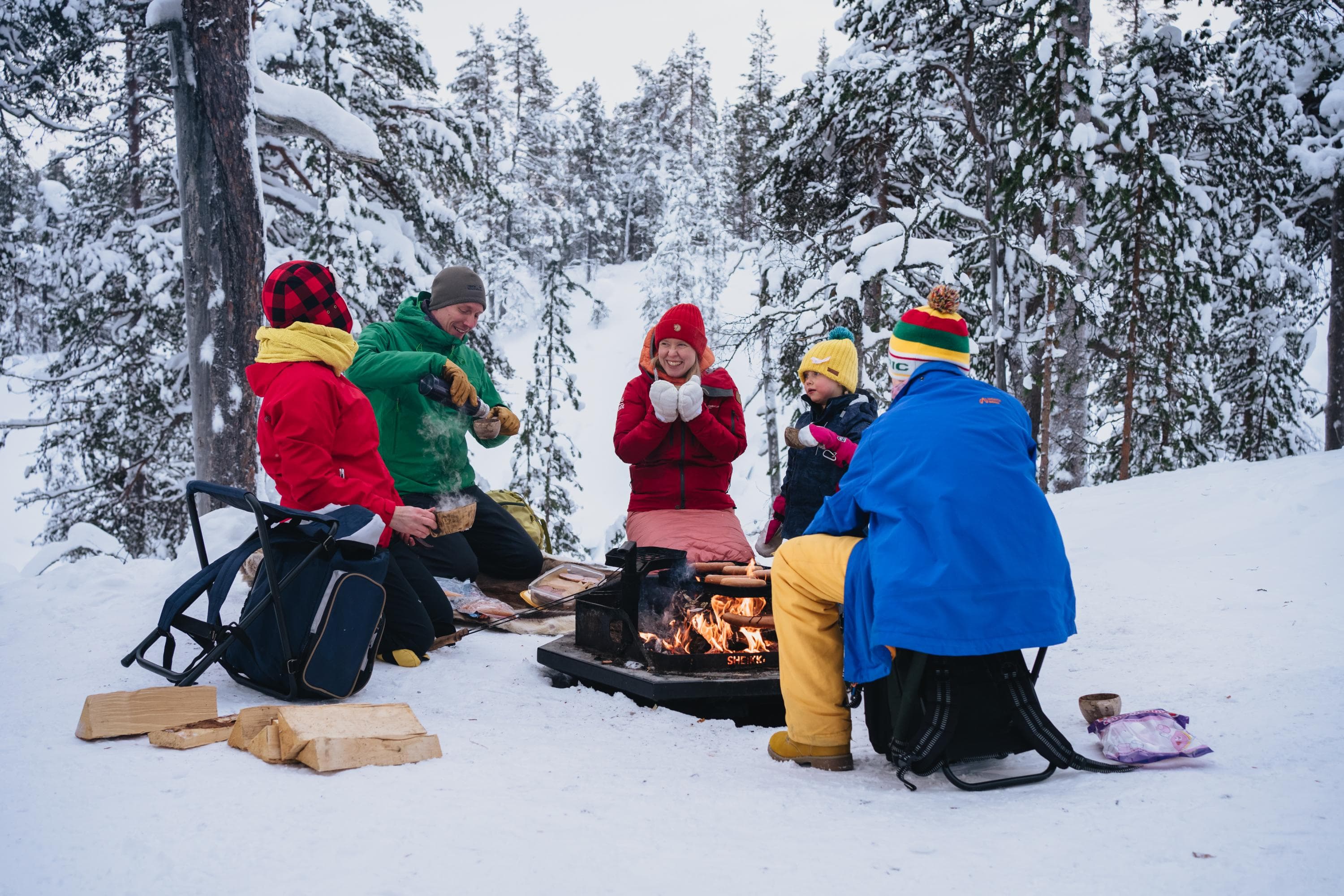Bild: Jaakko Posti Ihmiset istuvat nuotiotulen äärellä. Taustalla luminen metsämaisema.