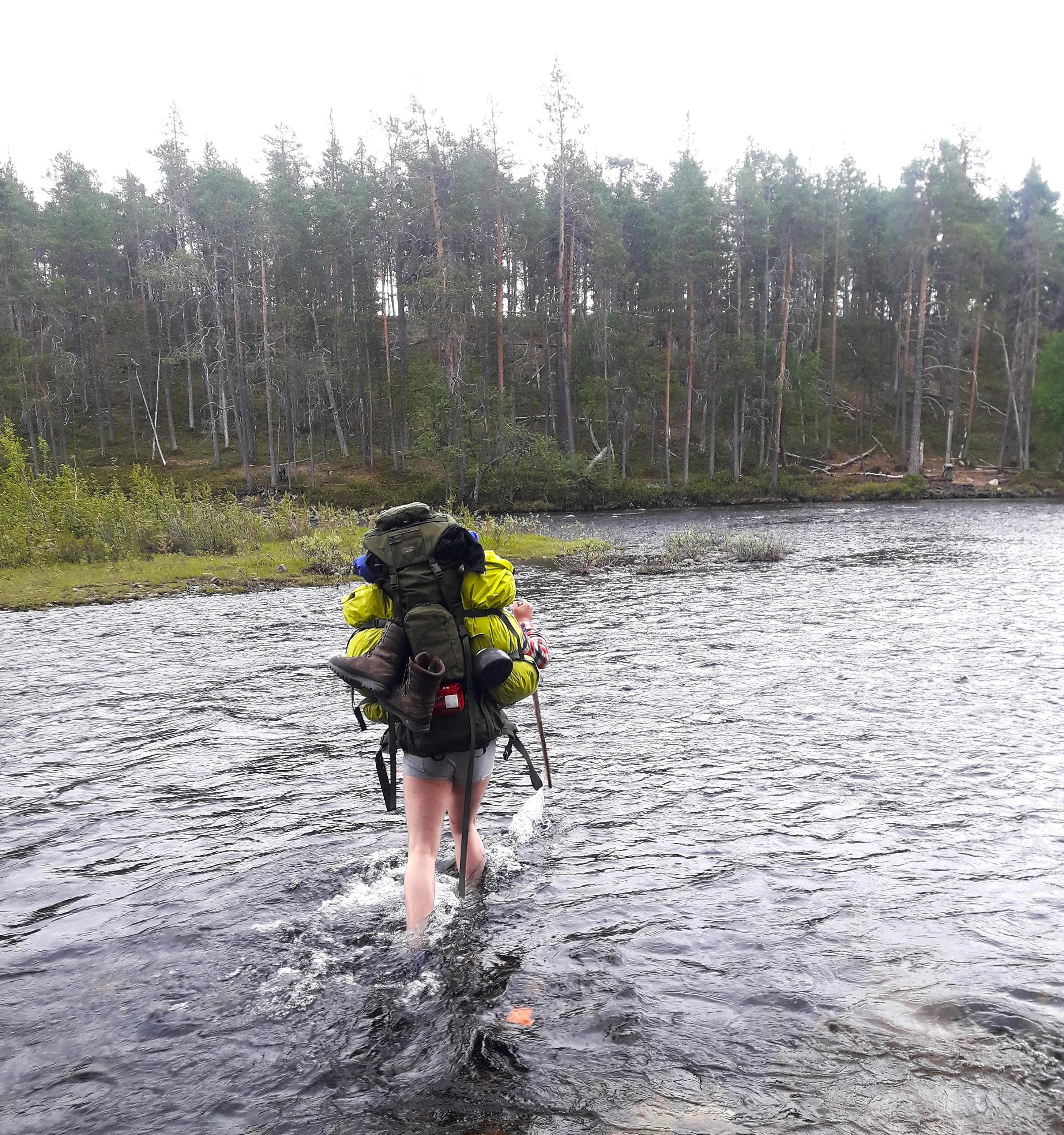Image: Kristiina Aikio A hiker wades across the river barefoot. He has a large backpack on his back.