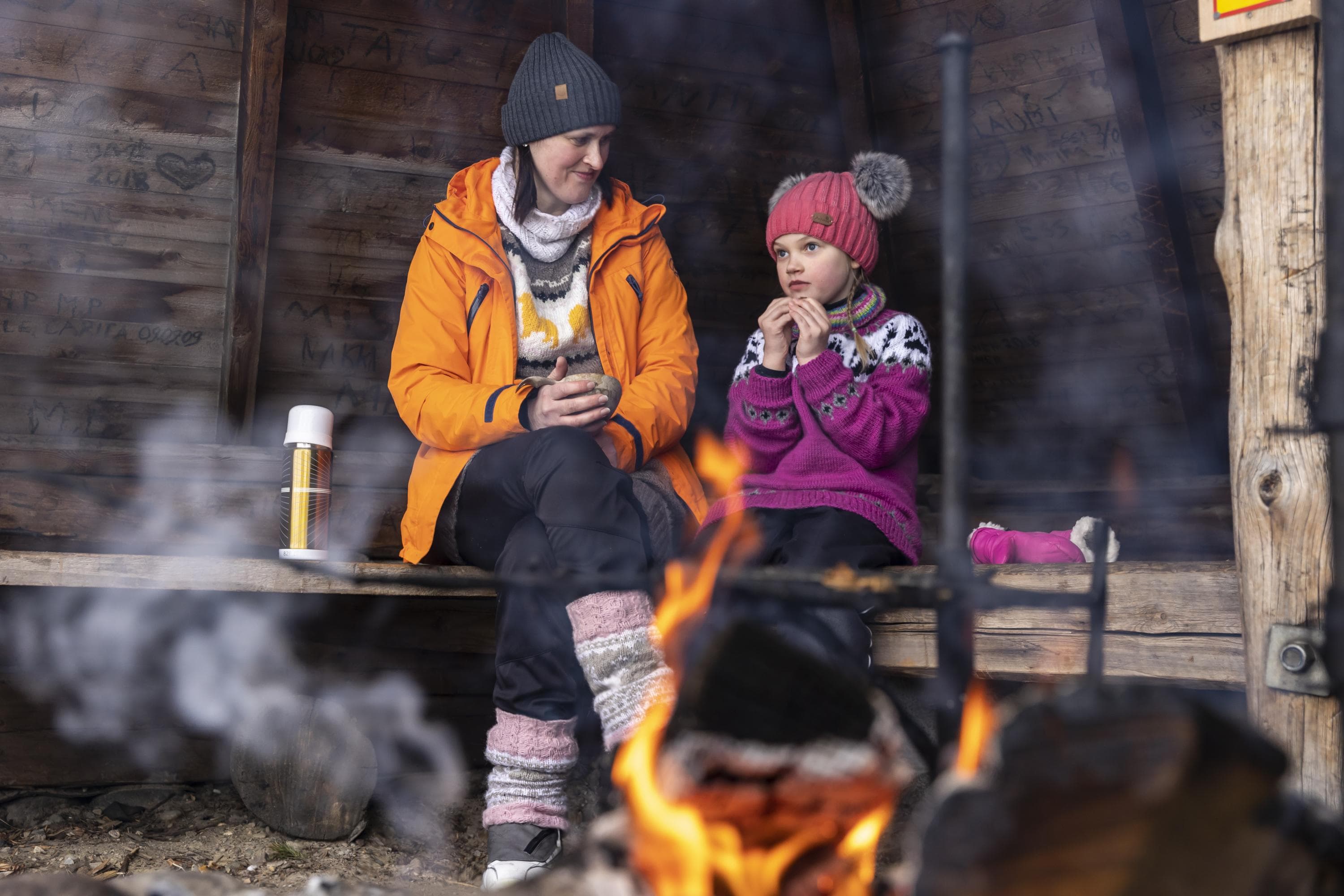 Image: Terhi Tuovinen A mother with a child taking a snack break at a lean-to shelter, wearing winter clothing.