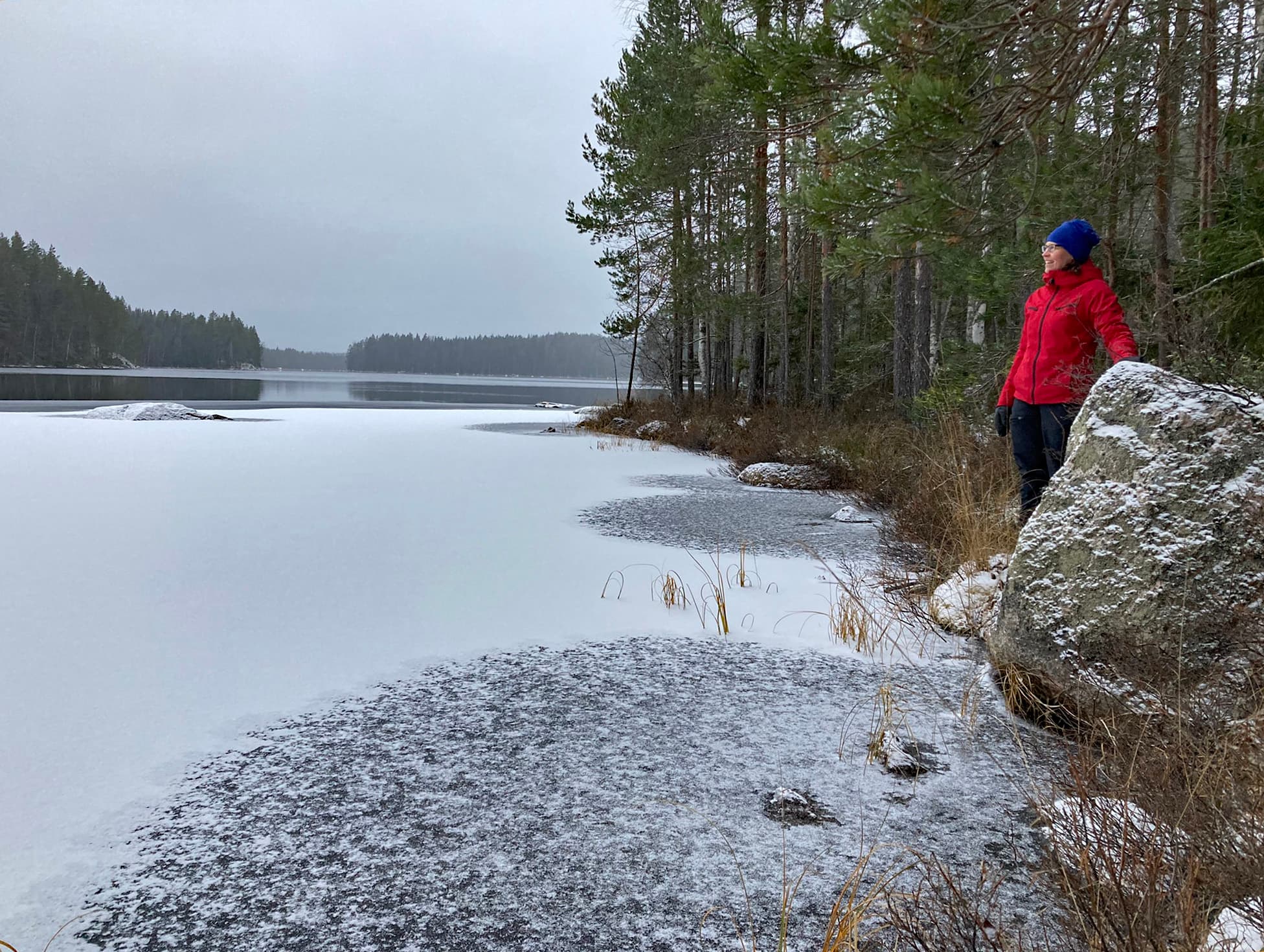 Image: Henri Solismaa A person stands next to a large rock on the shore, looking out over the lake. There is a thin layer of ice on the lake's surface near the shore, while the farther parts of the lake remain unfrozen.