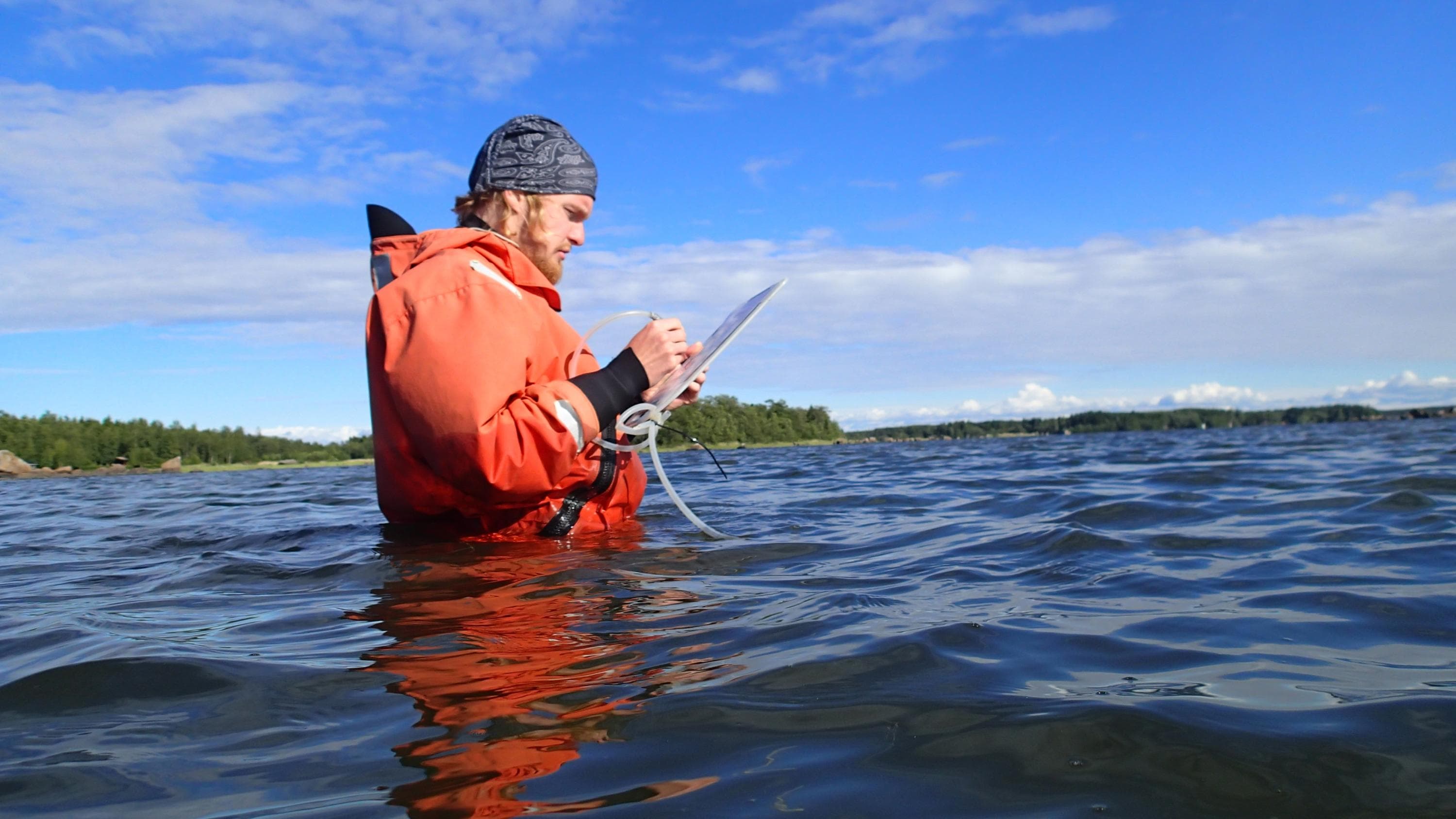 Kuva: Helmi Mentula Työntekijä on vyötäröä myöten vedessä ja kirjoittaa muistiinpanoa alustalle.