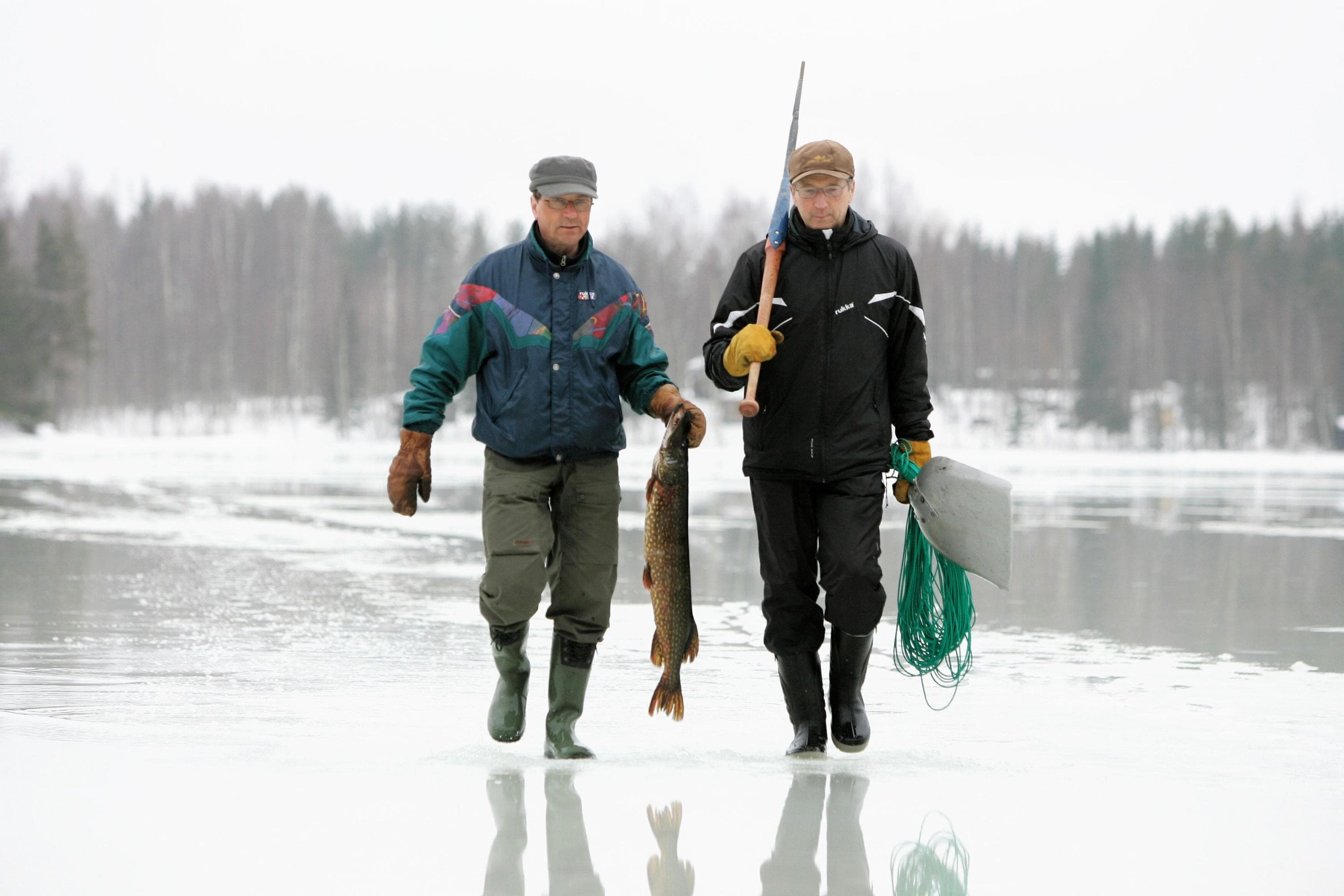 Image: Jere Malinen Two people walking on wet ice. One carries a large fish, and the other carries equipment used for winter net fishing.
