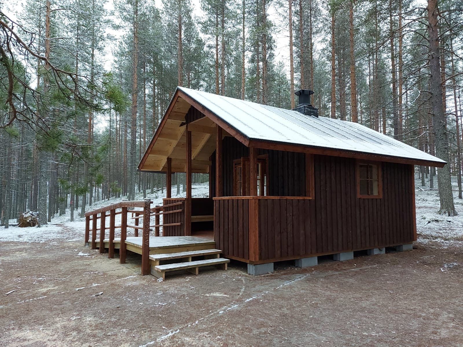 Image: Heidi Kontiokari A small brownish cabin at the edge of a pine forest. A light dusting of snow has fallen on the cabin's roof and the forest floor.