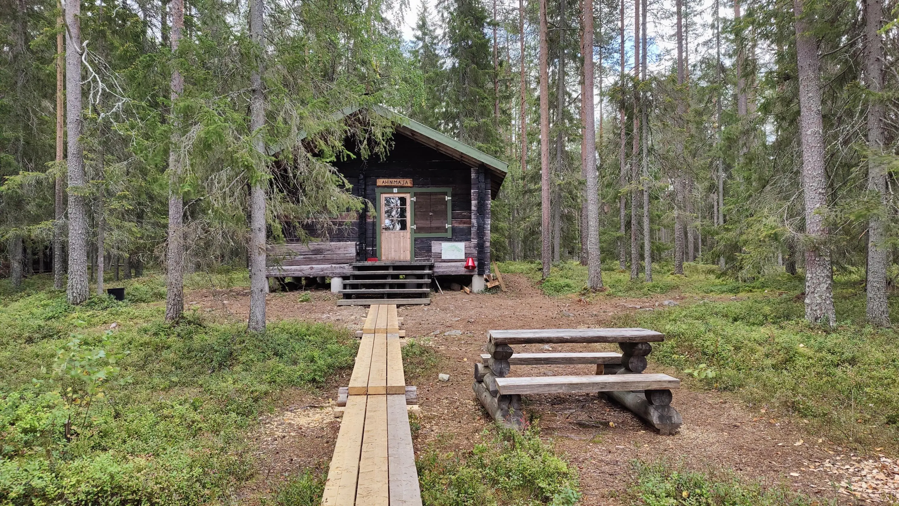 Bild: Ulla Matturi Spångar leder till en timmerstuga mitt i skogen. Till höger om spångarna finns ett picnic bord.