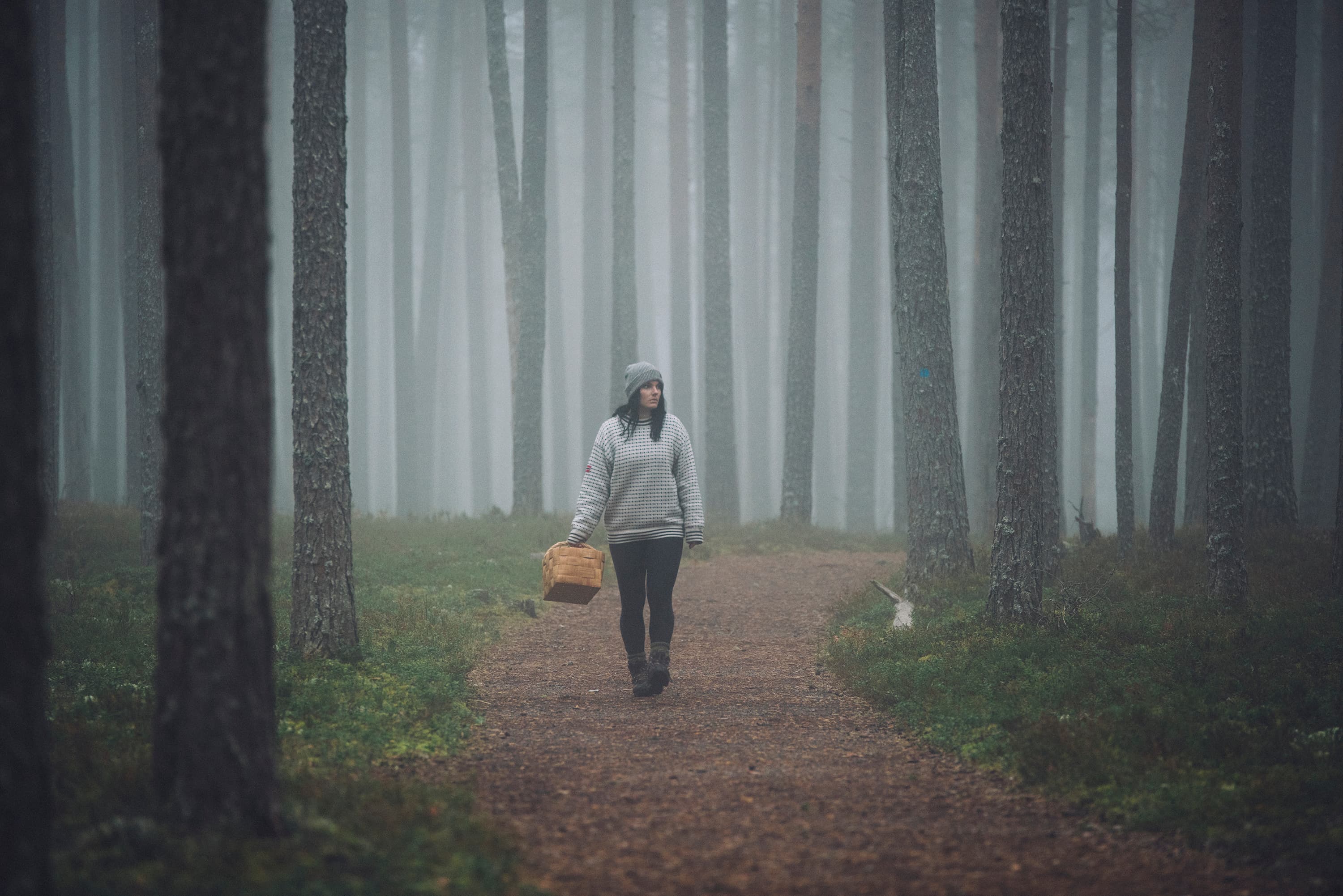 Kuva: Eeva Mäkinen Nainen kävelee korin kanssa polulla sumuisen mäntymetsän keskellä.