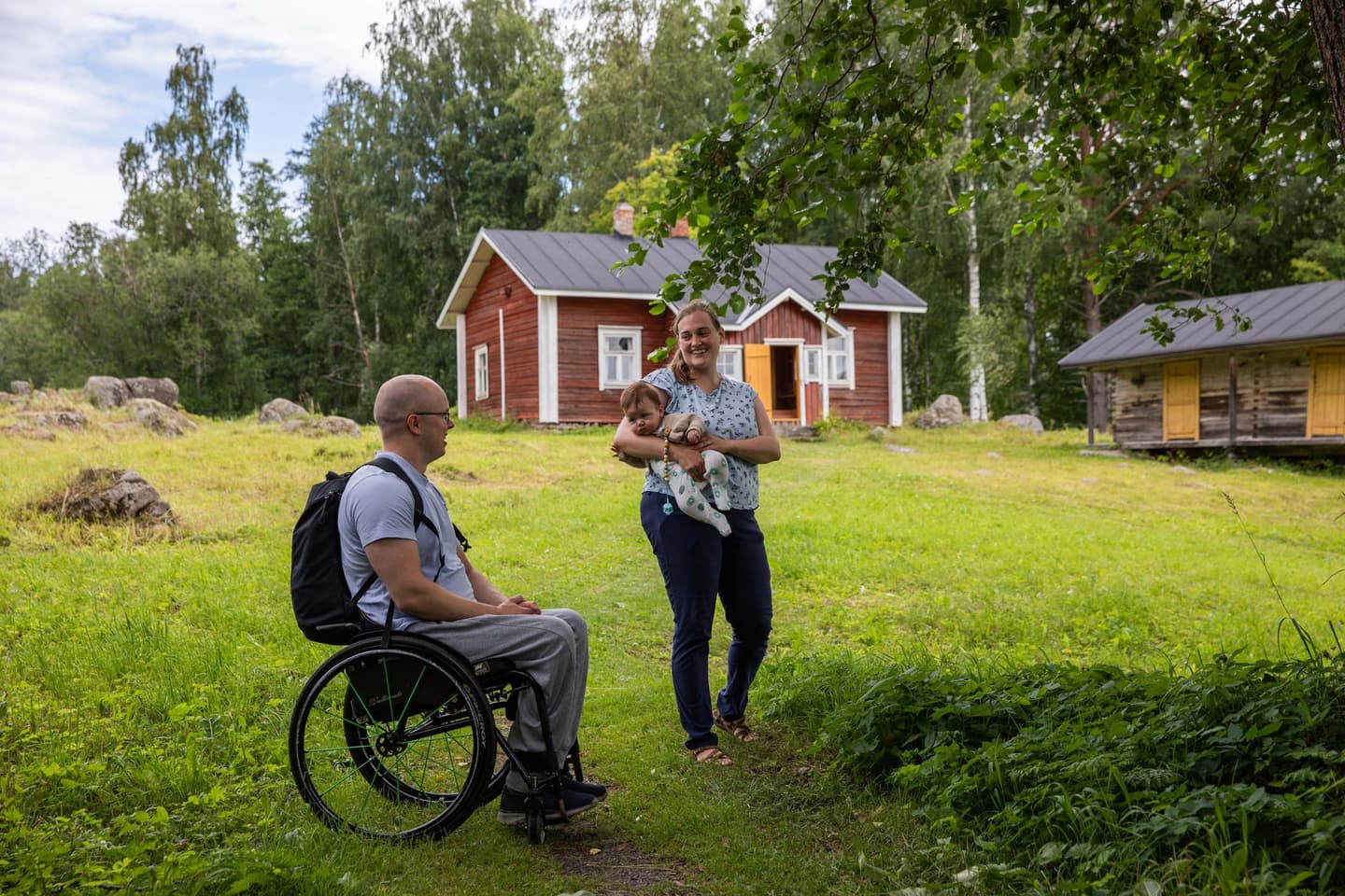Image: Saara Lavi Two hikers look at each other smiling. One is sitting in a wheelchair and the other is holding a baby. Wooden buildings in the background.