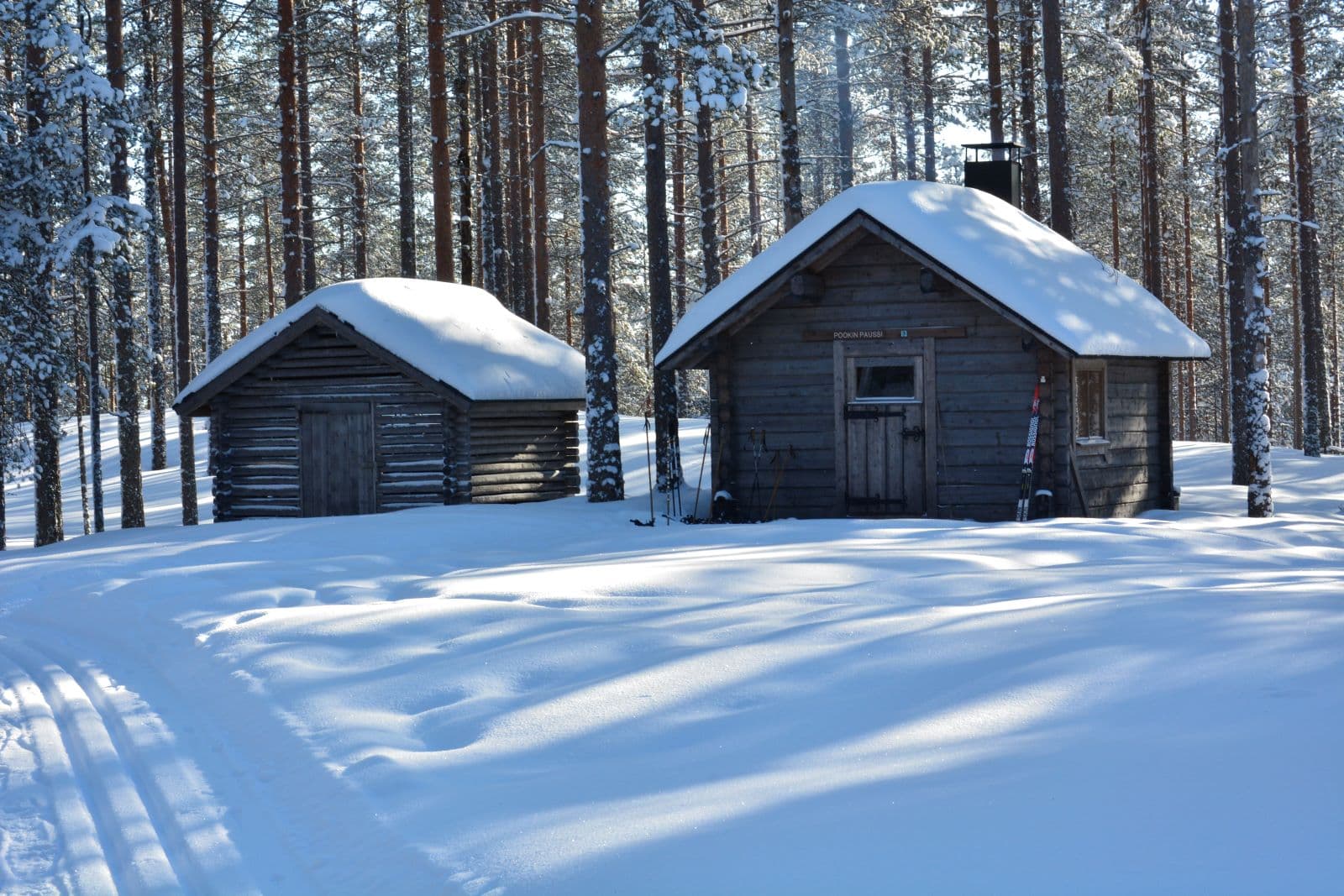 Kuva: Saara Airaksinen Kaksi harmaata mökkiä hiihtoladun varrella. Maa ja mökkien katot ovat lumiset.