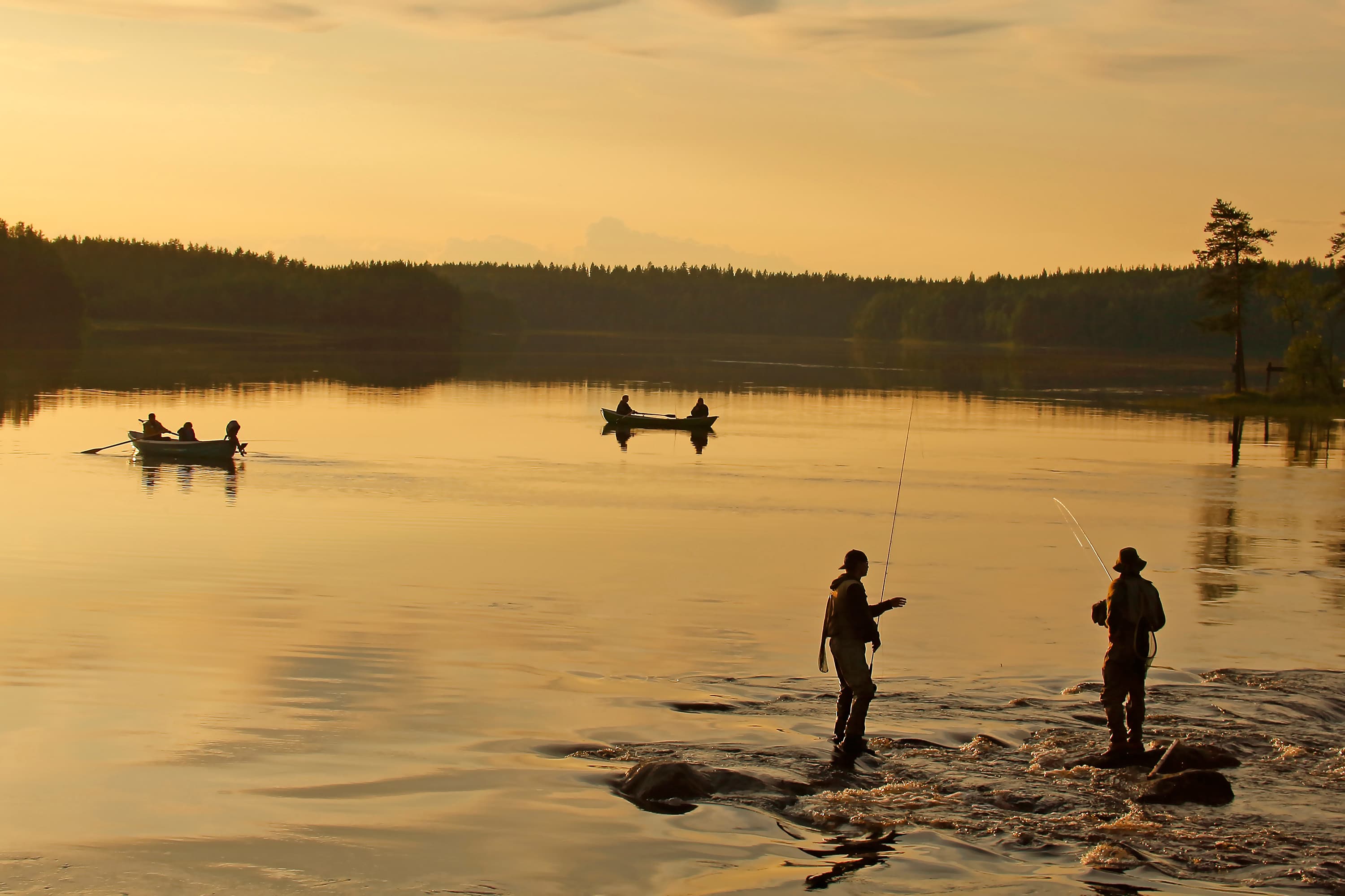 Kuva: Ismo Pekkarinen Järvimaisemassa kaksi henkilöä seisoo kuvan etuosassa kalassa koskipaikassa ja järvellä soutelee kaksi venettä.