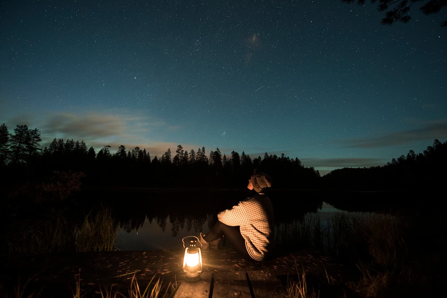 Bild: Eeva Mäkinen En person sitter på en brygga med en lykta och blickar upp mot stjärnhimlen. Den mörka siluetten av skogen på andra sidan stranden speglas i vattenytan.
