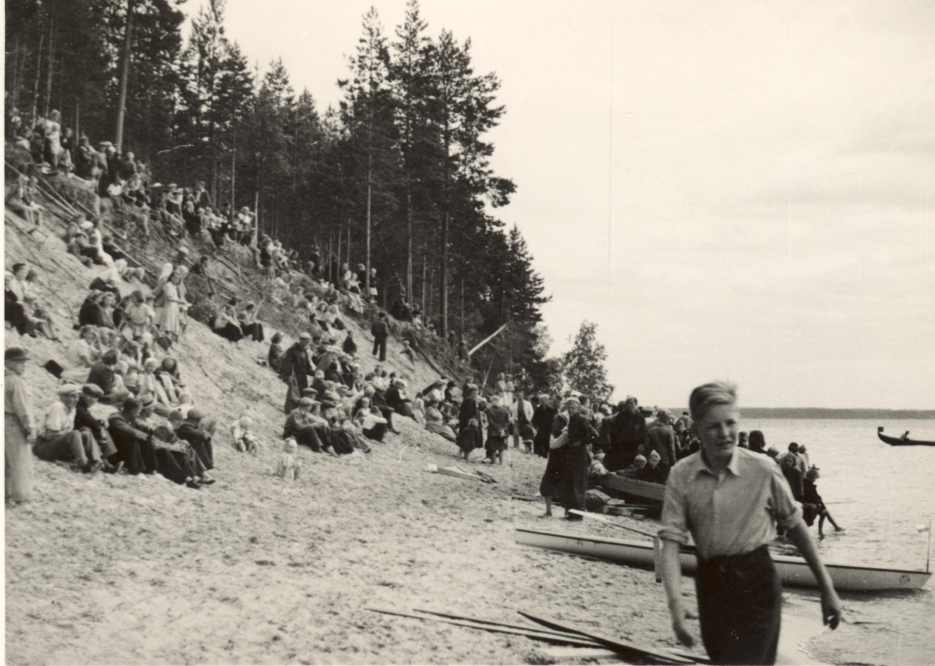 Image: Okänd / Kainuu museo On the left, a sand dune with many people sitting on it. On the right side, a lake.