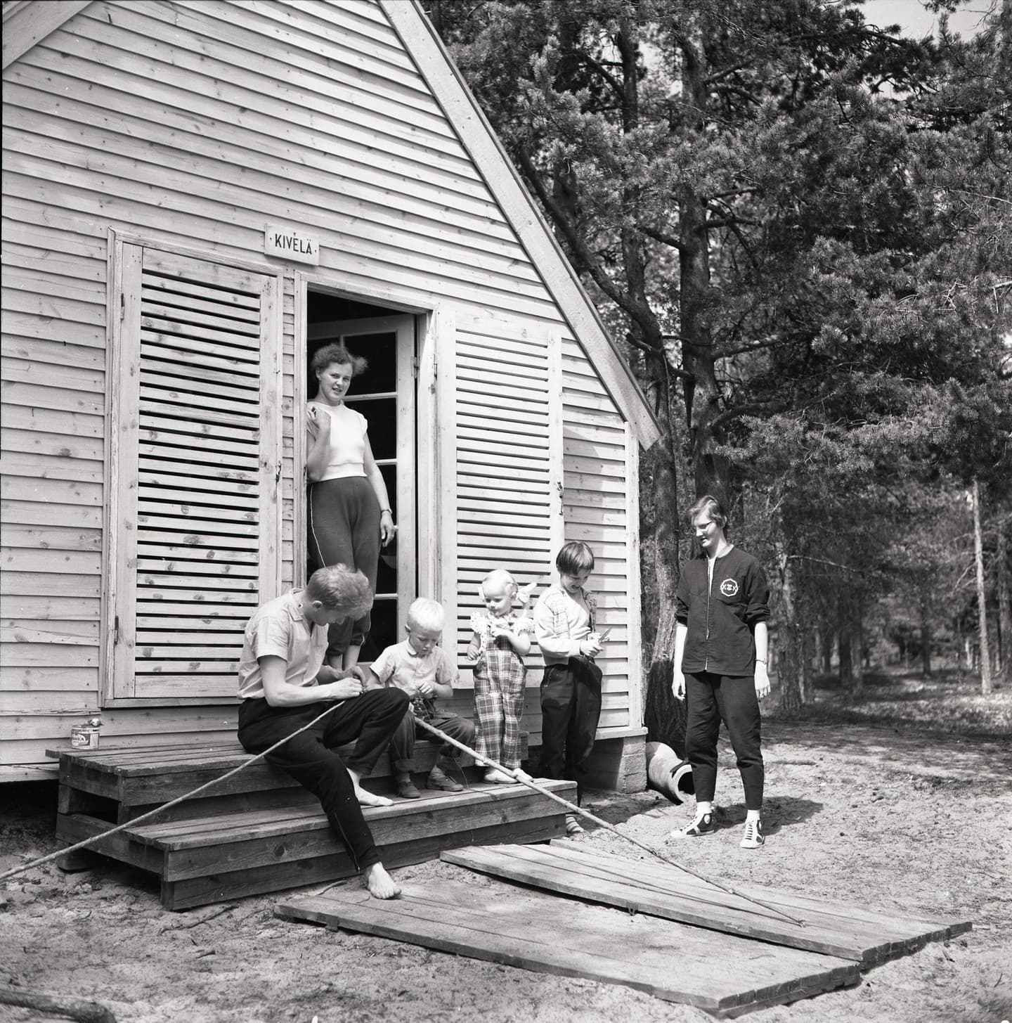 Image: Okänd / Kainuu museo On the left side, a small holiday cottage with people in front of it. On the right side, a forest.
